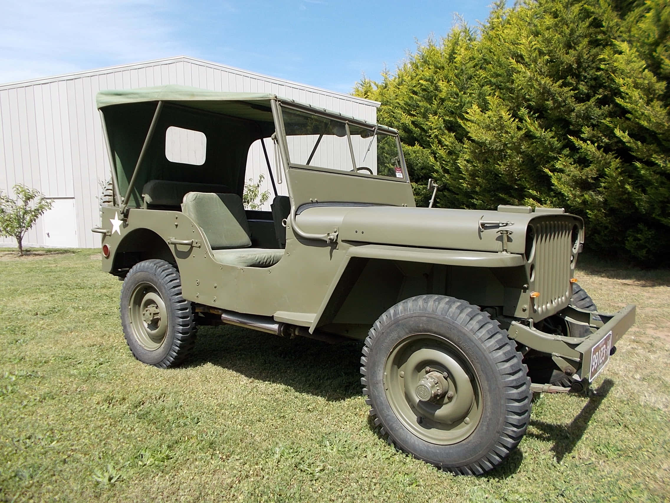 Jeep Willys 桌布 壁紙 — 一輛經典的橄欖綠色軍用吉普車停泊在草地上，旁邊是白色建築物和茂密的綠樹，天空清澈湛藍。