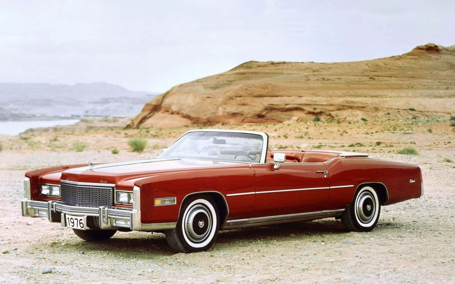 Cadillac Eldorado 桌布 壁紙 — 一輛經典紅色1976年凱迪拉克開篷車停泊在碎石沙漠道路上，背景有岩石山丘和遠處的水面。