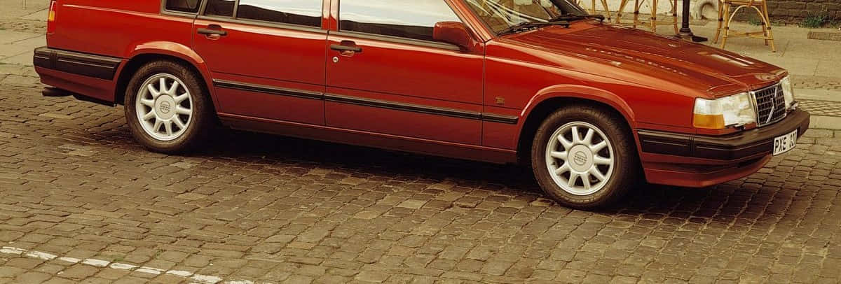 Volvo 940 桌布 壁紙 — 一輛經典紅色旅行車停泊在鵝卵石街道上，背景有木椅。