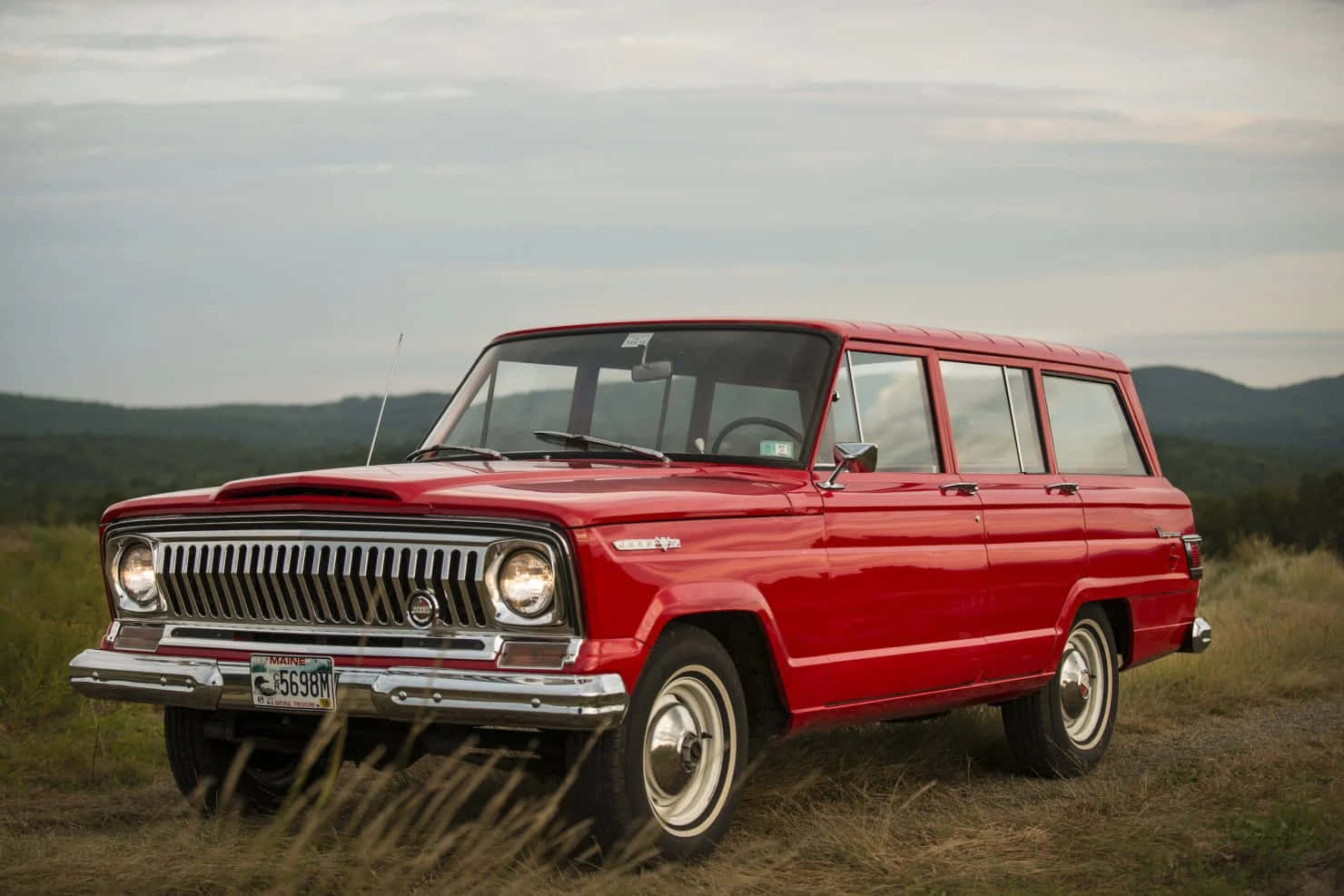 Jeep Wagoneer 桌布 壁紙 — 一輛經典紅色旅行車停泊在草地地形上，背景是起伏的丘陵，天空多雲。
