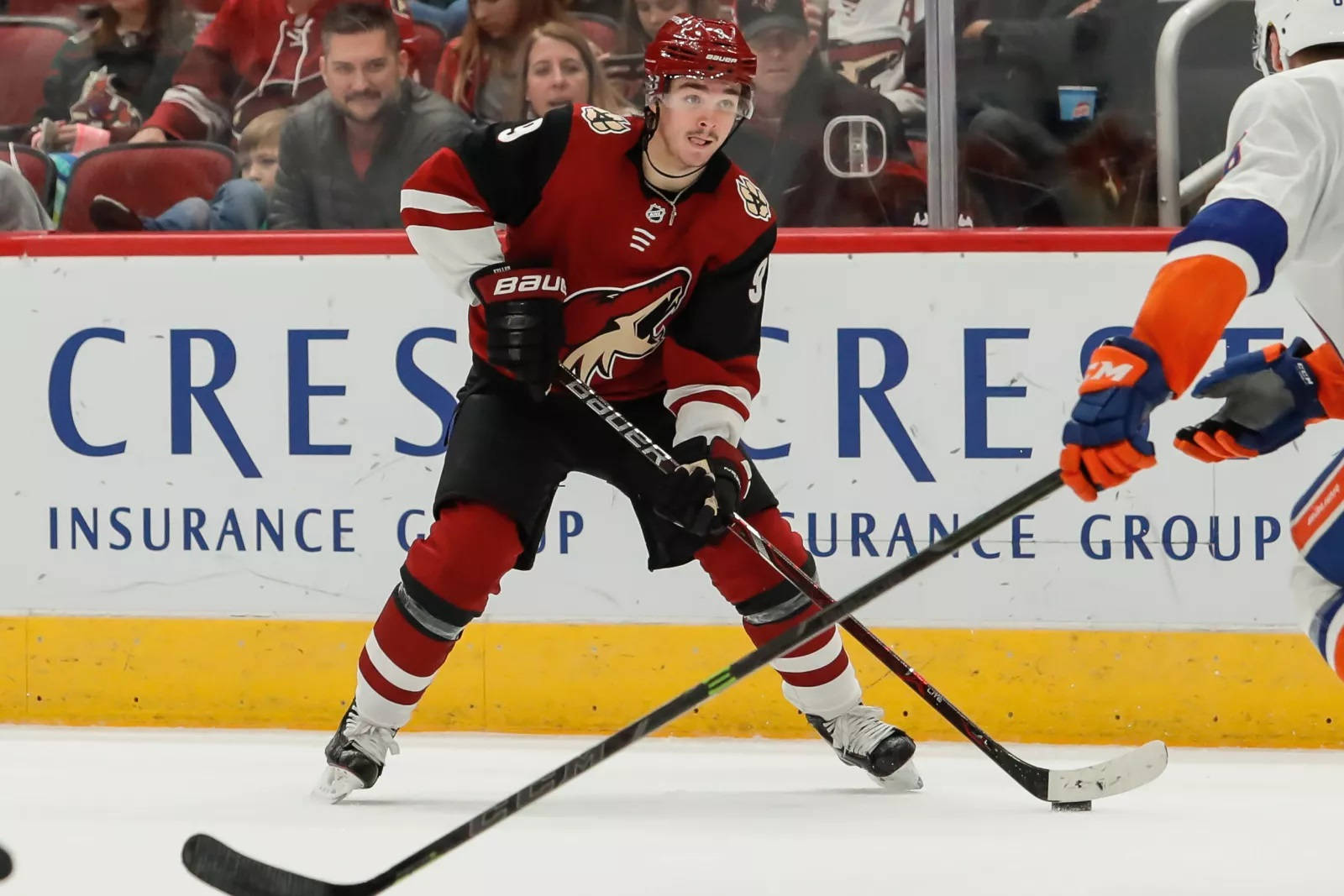 Clayton Keller 2018 NHL賽事 壁紙 — 一名身穿紅黑裝備的冰球運動員在比賽中，背景有觀眾，在冰上激烈滑行，球棍上帶著冰球。