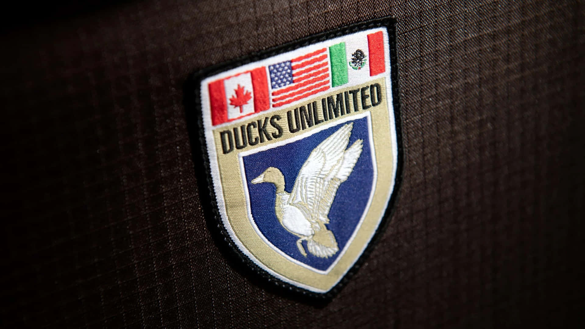  壁紙 — 近距離拍攝Ducks Unlimited徽章，上有飛鴨標誌及三面國旗，背景為深色布料。