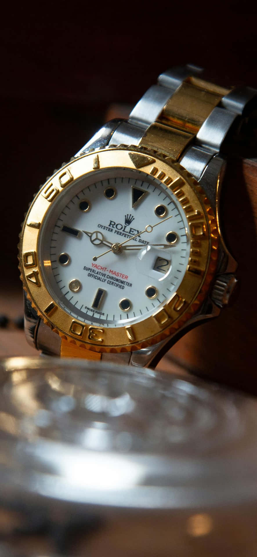  壁紙 — 金色和鋼製勞力士Yacht-Master手錶的特寫，放在木質表面上，旁邊有一個模糊的玻璃杯。