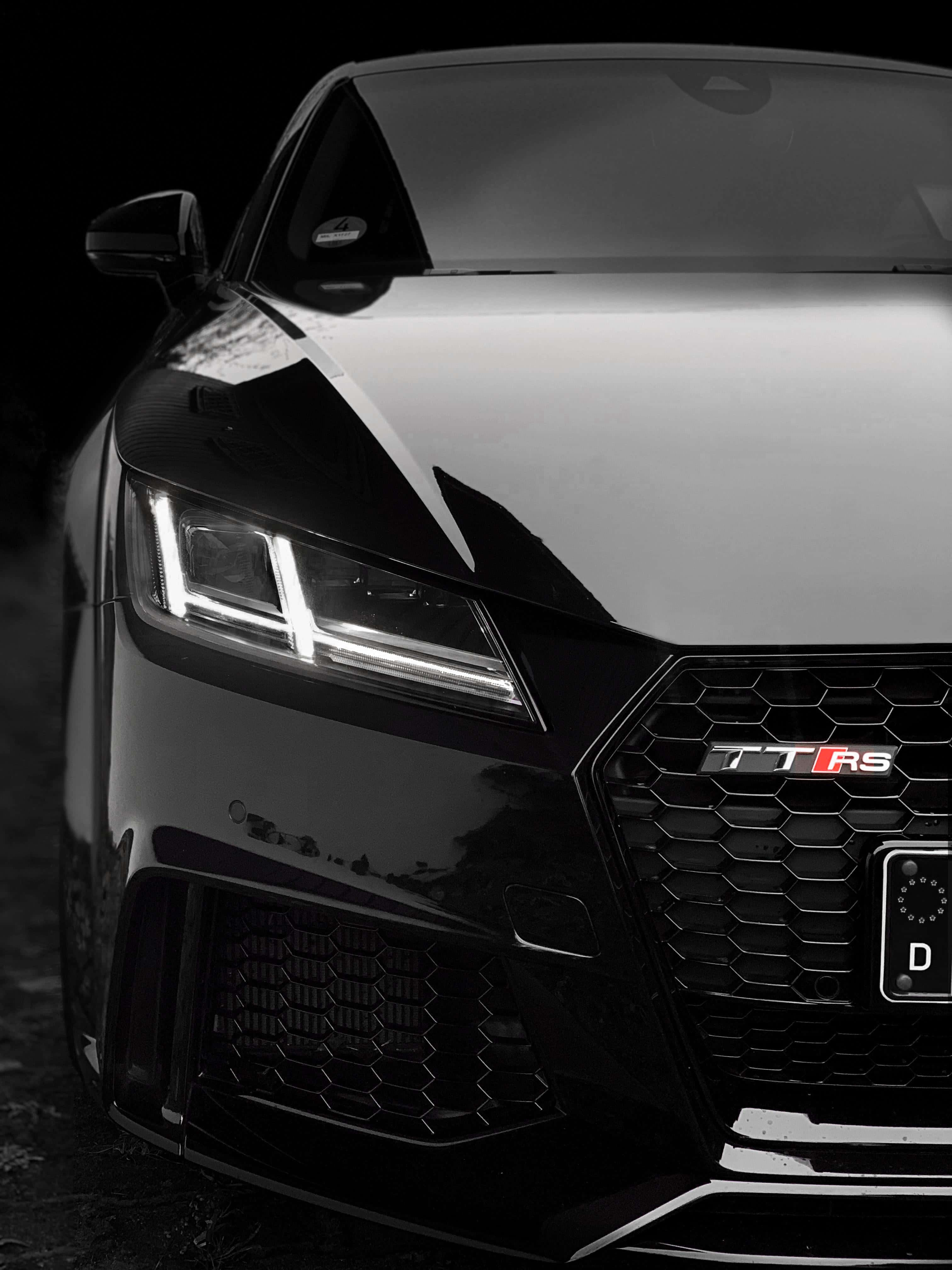 Audi Tt Rs 桌布 壁紙 — 黑色奧迪 TT RS 跑車特寫，配備發光頭燈和六角形鬼面罩，背景漆黑。