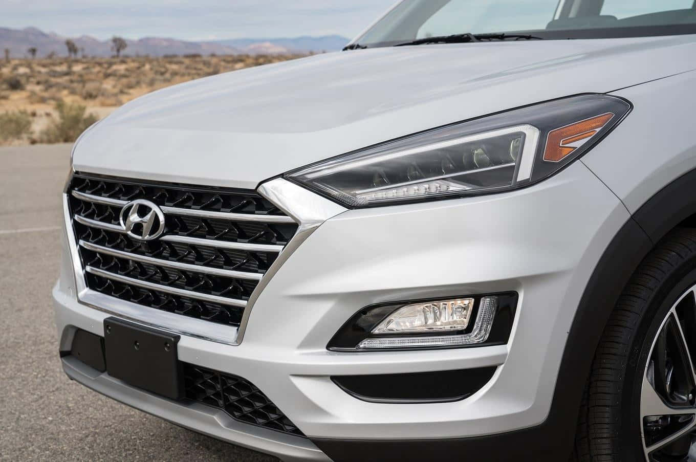 Hyundai Tucson 桌布 壁紙 — 一輛時尚白色現代SUV的前格柵和頭燈特寫，停在沙漠公路上，遠處有山脈。