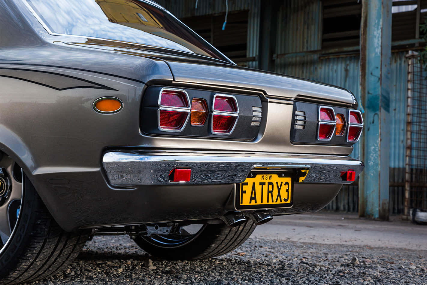 Mazda Rx-3 桌布 壁紙 — 復古灰色轎車尾部特寫，帶有鍍鉻保險桿和黃色車牌，停在碎石地上，靠近生鏽的金屬建築物。