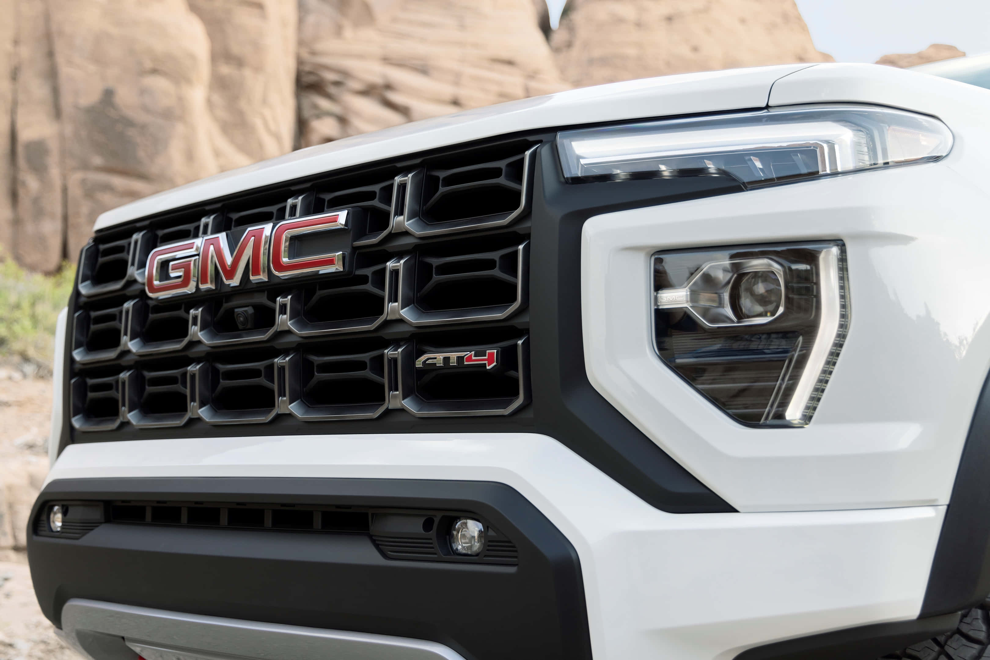 Gmc Canyon 桌布 壁紙 — 白色GMC AT4卡車前格柵和頭燈在岩石沙漠地形中的特寫。
