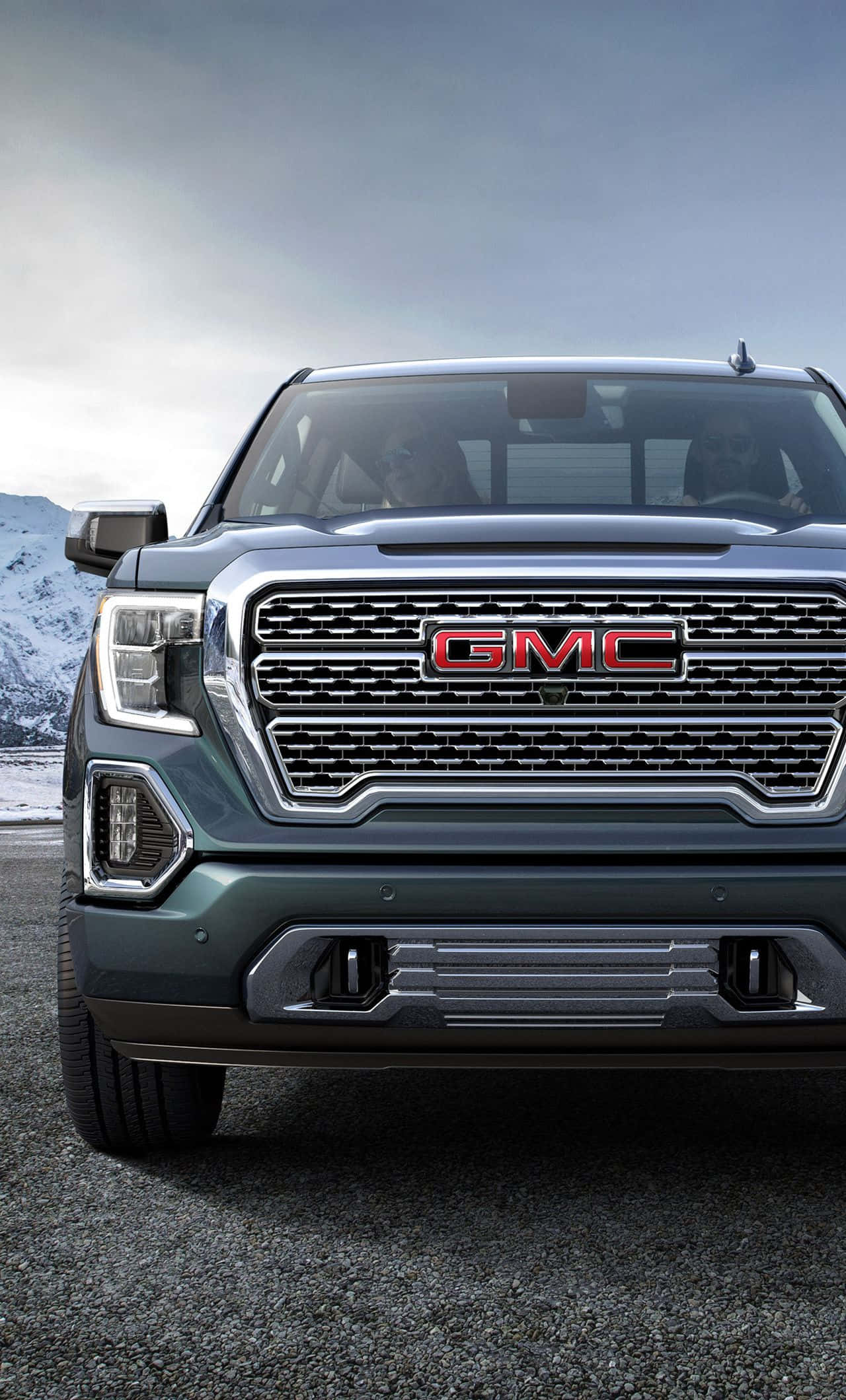 Gmc Yukon 桌布 壁紙 — 特寫鏡頭：深綠色GMC貨車的前面格柵，帶有紅色標誌，背景是陰天下的雪山。
