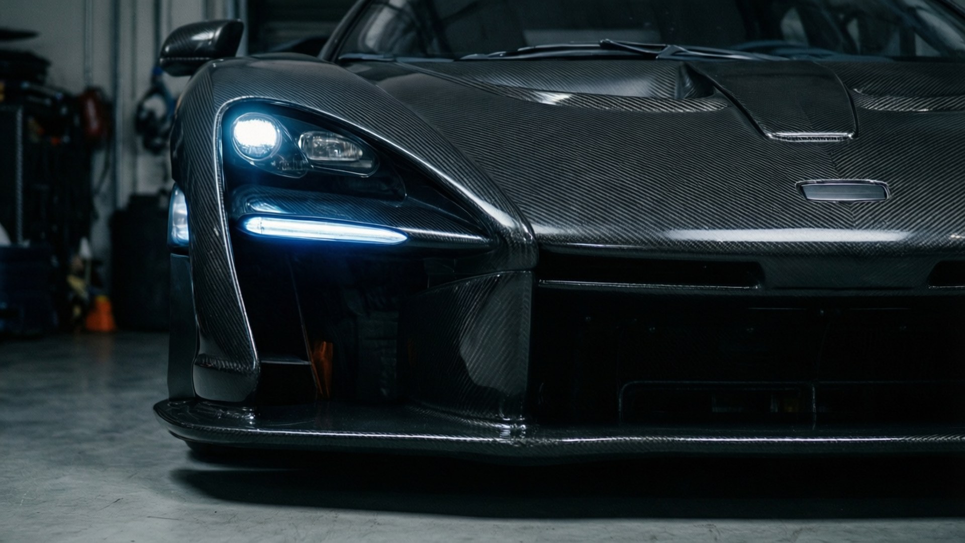 Mclaren Senna 桌布 壁紙 — 一輛時尚黑色碳纖維跑車在昏暗車庫中亮著車頭燈的特寫。
