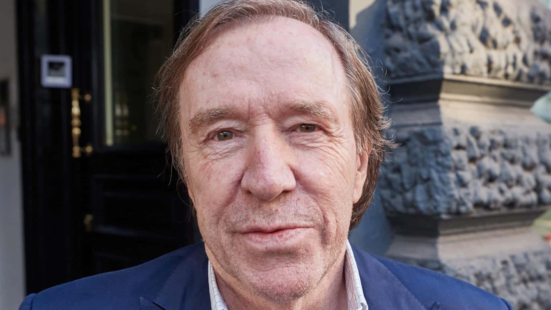  壁紙 — 中年男子嘅特寫肖像，佢有棕色頭髮、著藍色西裝外套，企喺室外嘅石紀念碑同黑色門附近。