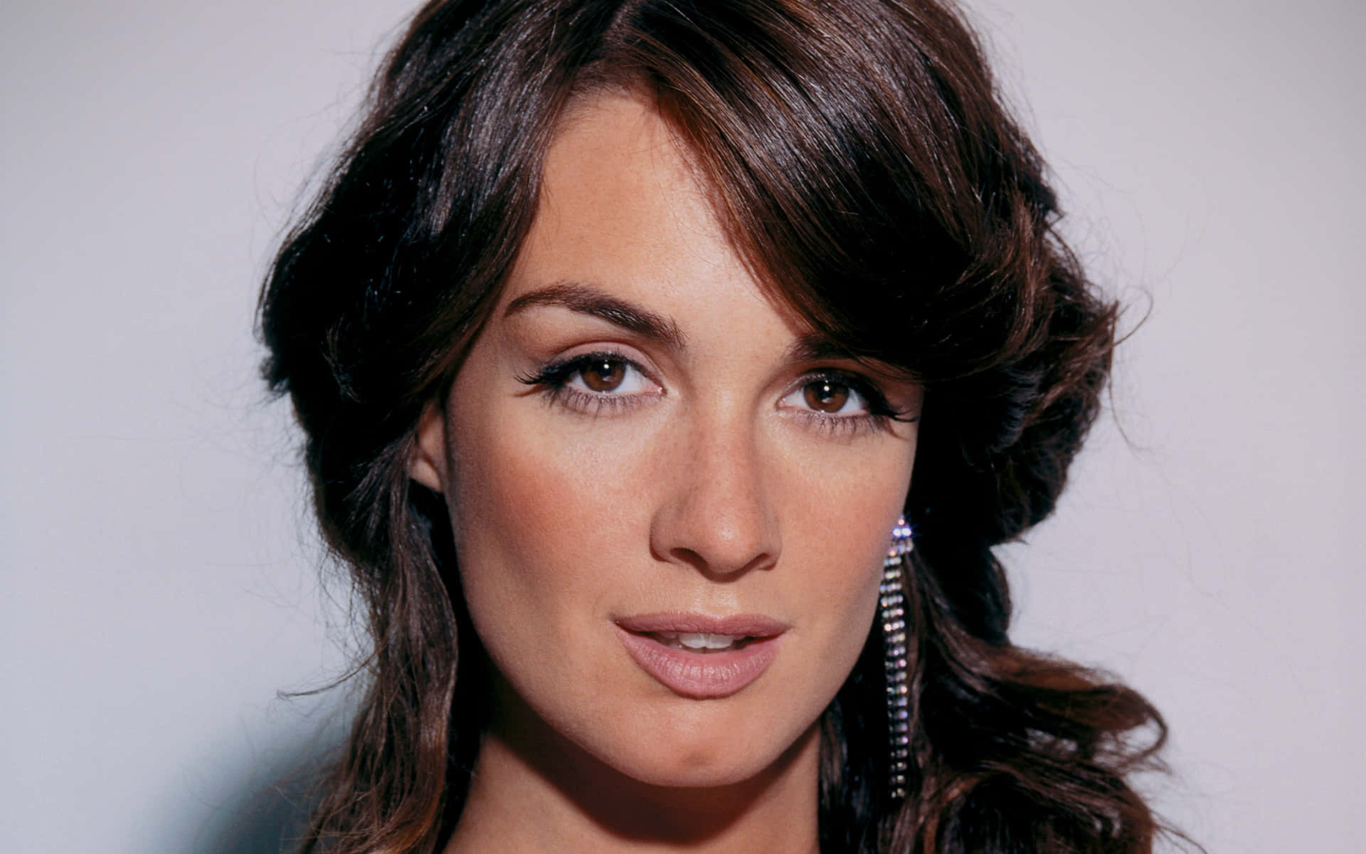 Paz Vega 桌布 壁紙 — 一位深色波浪髮女性戴著閃亮垂墜耳環的特寫肖像，背景為純淺灰色。