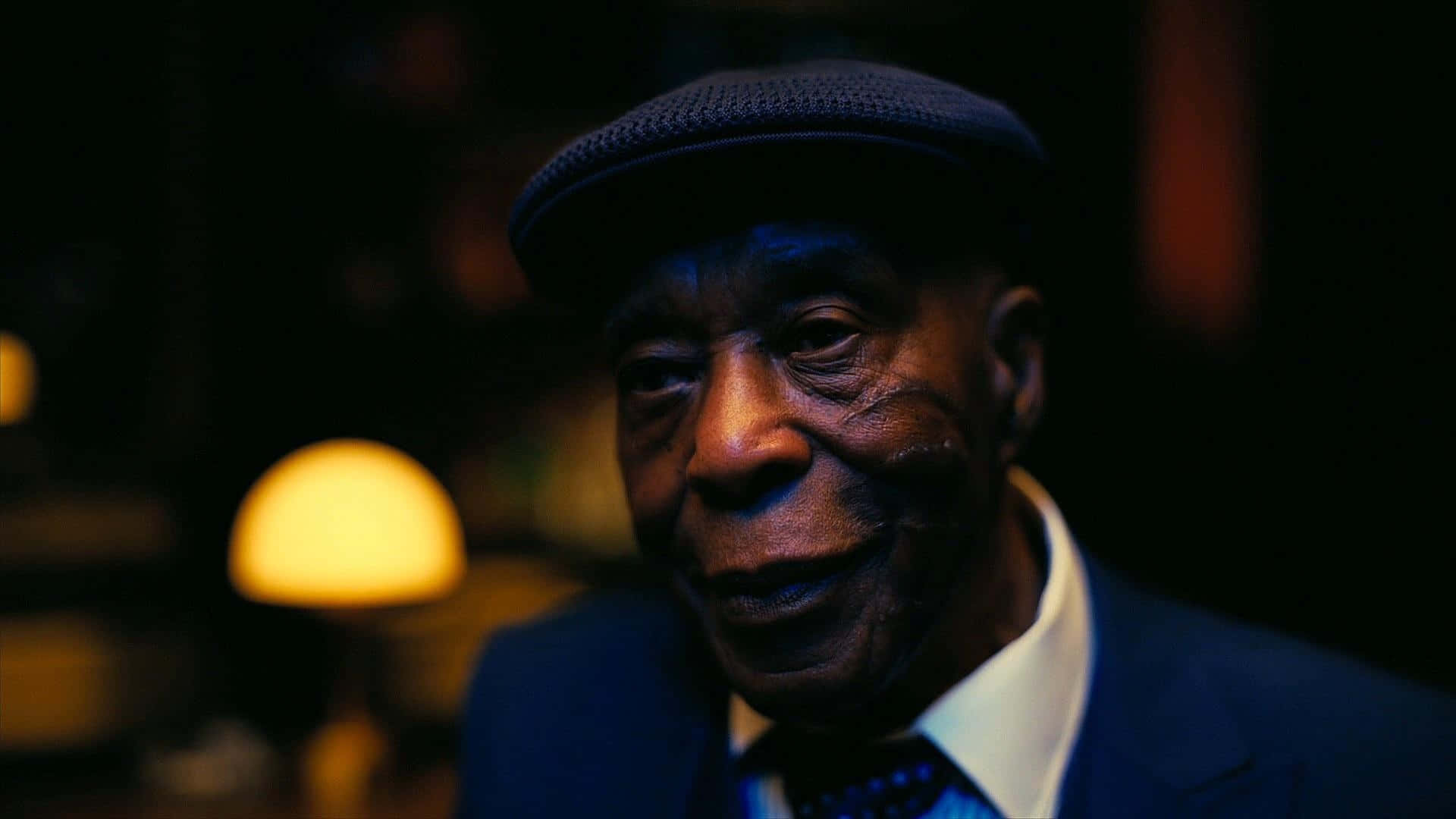  壁紙 — 一位戴著深色帽子和西裝的年長黑人男子的特寫肖像，在昏暗的環境燈光下，背景中有一盞發光的燈，他溫暖地微笑。