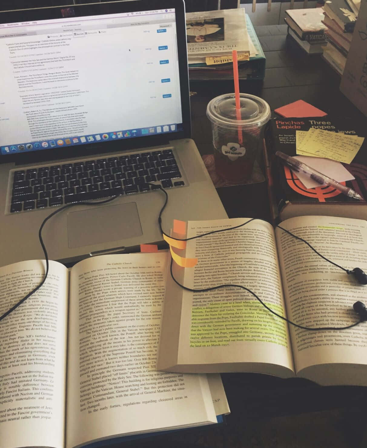  壁紙 — 一張雜亂的書桌，上面有一本打開的書、耳機、咖啡和一部顯示網頁的筆記型電腦，處於溫暖的室內燈光下。