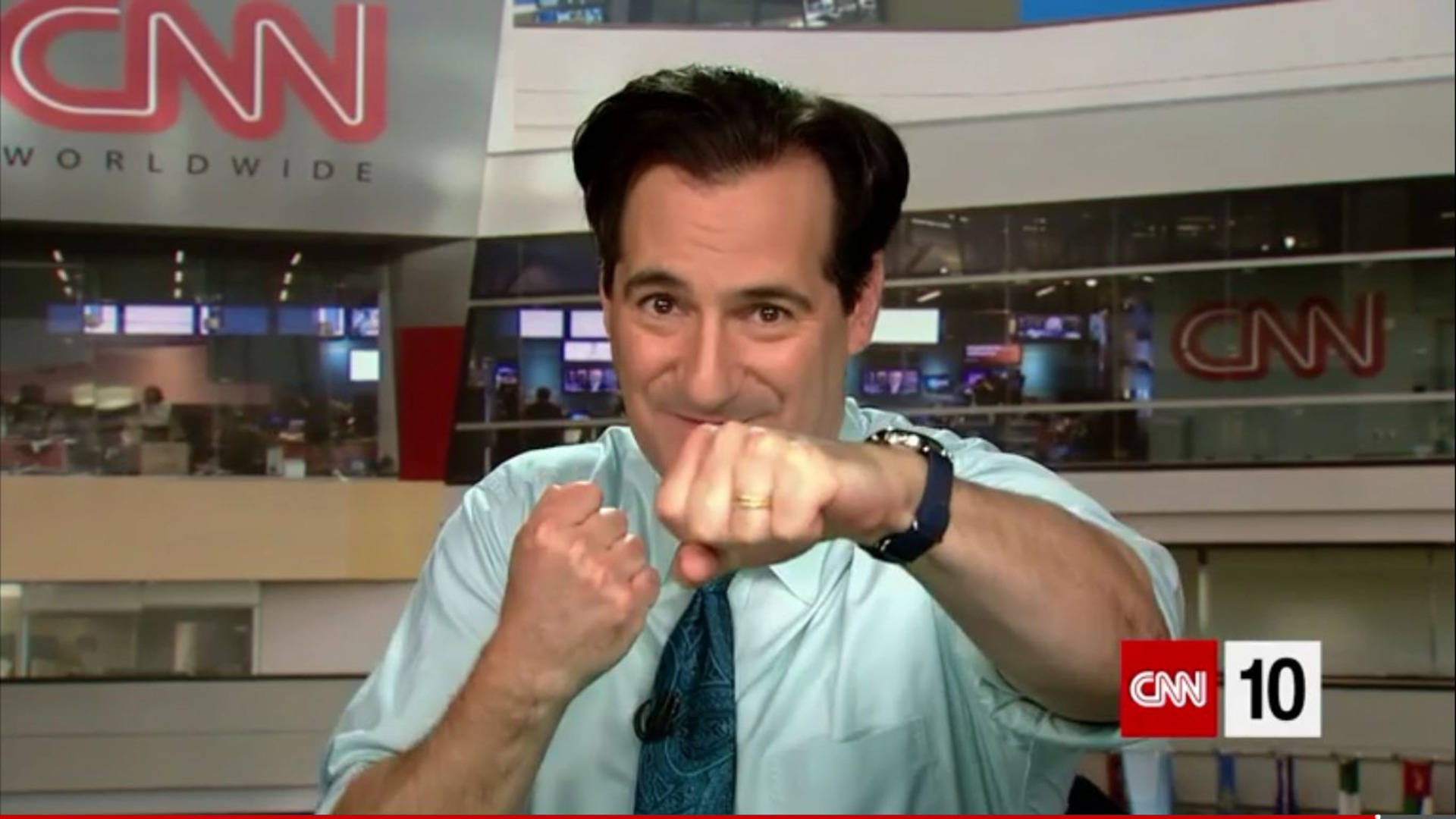 CNN 10 主持人卡爾·阿祖茲 壁紙 — CNN 錄影廠內一位微笑的新聞主播向鏡頭做出一個玩樂式的拳頭碰撞手勢。