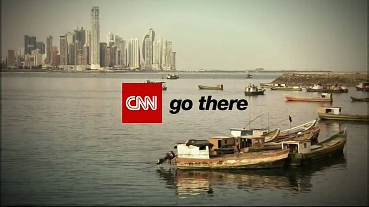 CNN Go There 巴拿馬 壁紙 — 飽經風霜的漁船漂浮在平靜的水面上，遠處朦朧中升起現代城市的天際線。