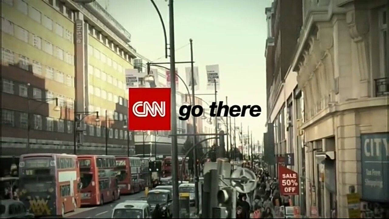 CNN Go There 紐約 壁紙 — 一條繁忙的城市街道，充滿紅色雙層巴士和行人，天空朦朧，帶有CNN go there標誌覆蓋層。