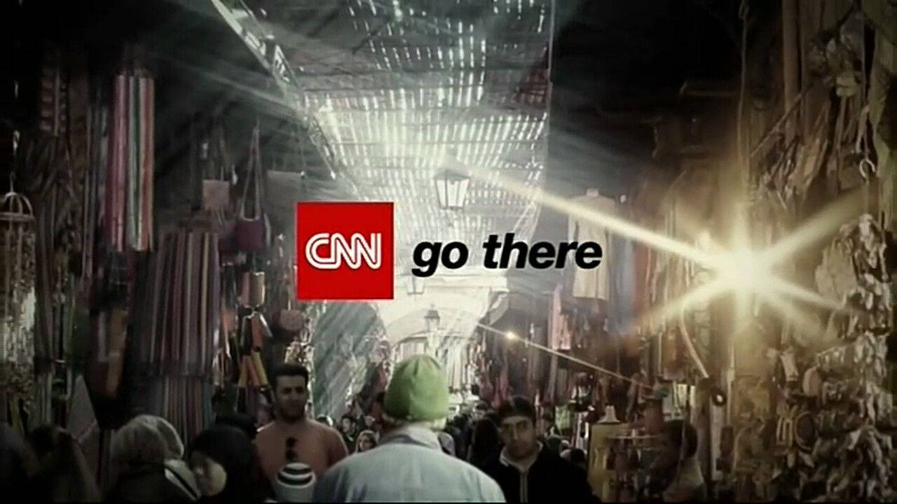 CNN Go There 馬拉喀什 壁紙 — 擁擠的市場小巷，掛滿紡織品，頭頂有明亮的陽光。