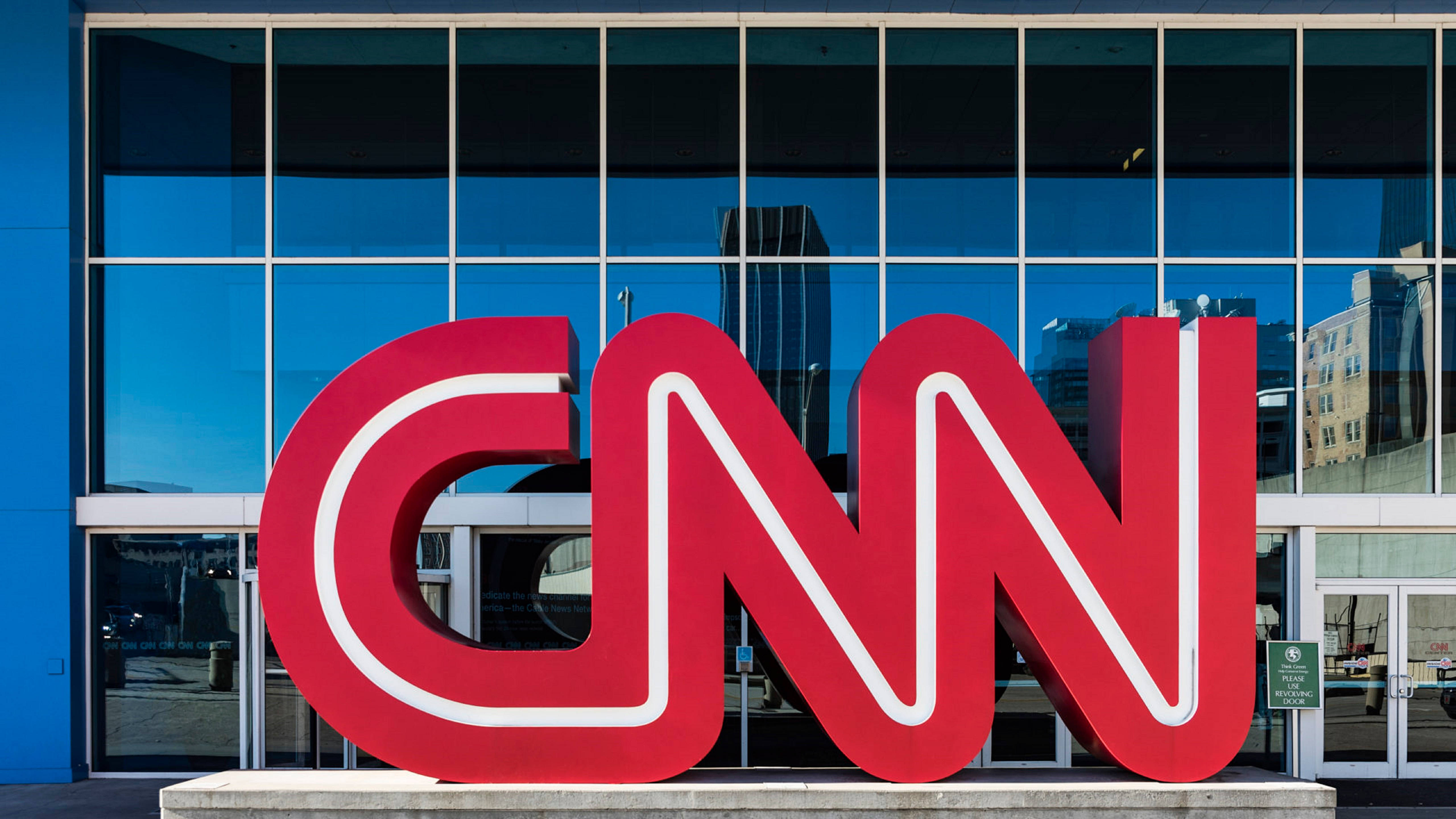CNN 亞特蘭大總部 壁紙 — 大型紅色CNN標誌醒目地矗立於現代玻璃建築前，建築物在明亮日光下反射城市摩天大樓。