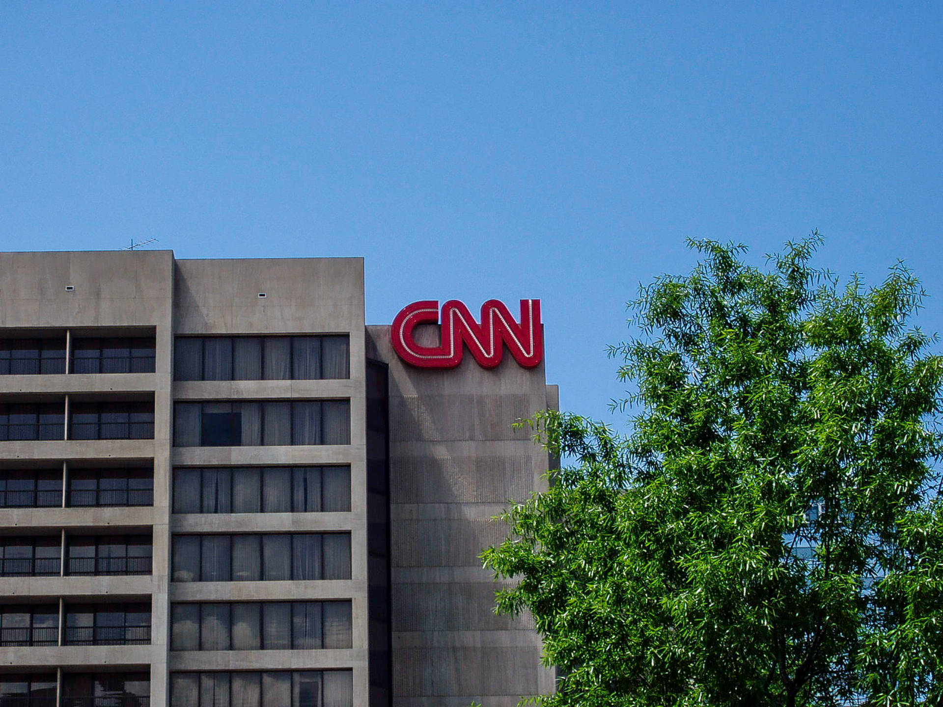 CNN總部大樓 壁紙 — 一座高聳的灰色建築物，上面有醒目的紅色CNN標誌，矗立在清澈的藍天下，部分被茂密的綠色樹枝環繞。