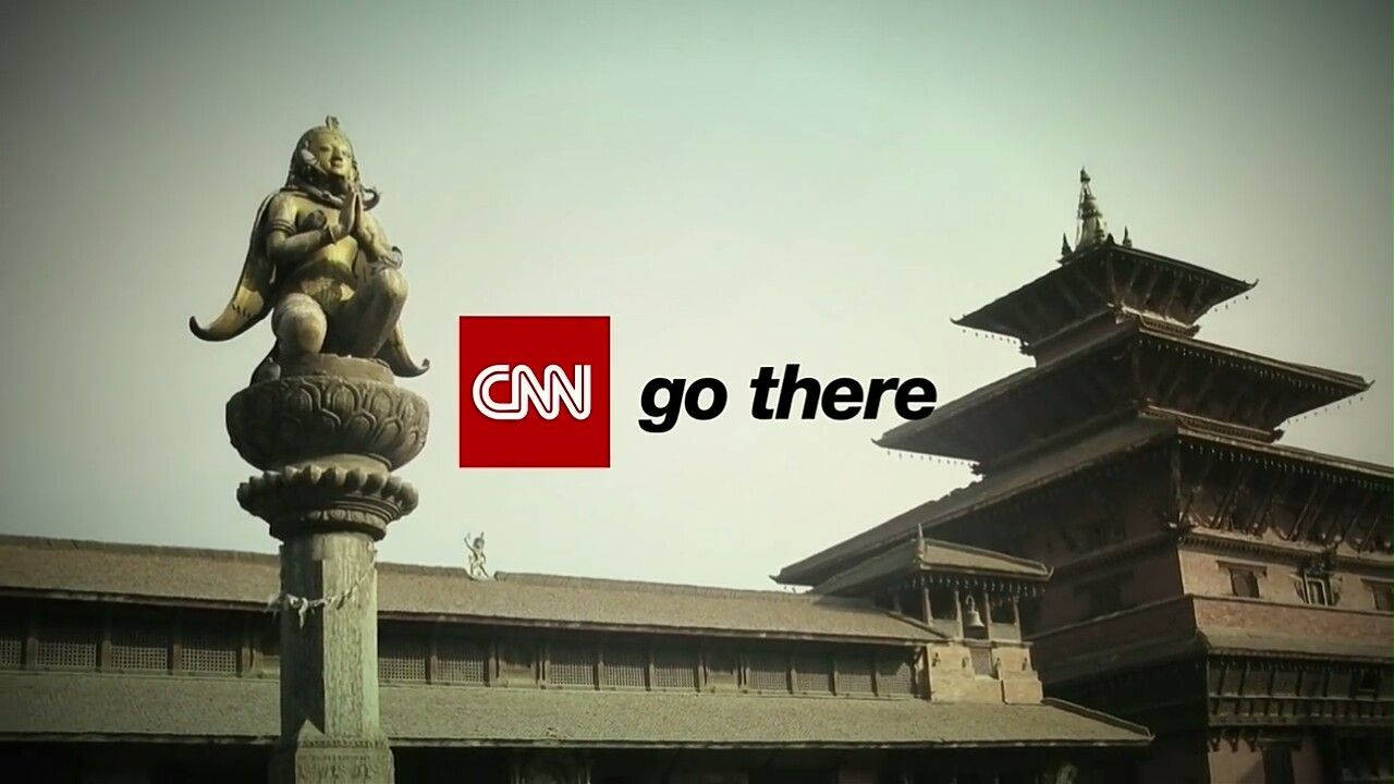 CNN記者於加德滿都報導 壁紙 — 一座神話人物的風化石像坐落於柱頂，旁邊是傳統多層寺廟，天空朦朧。