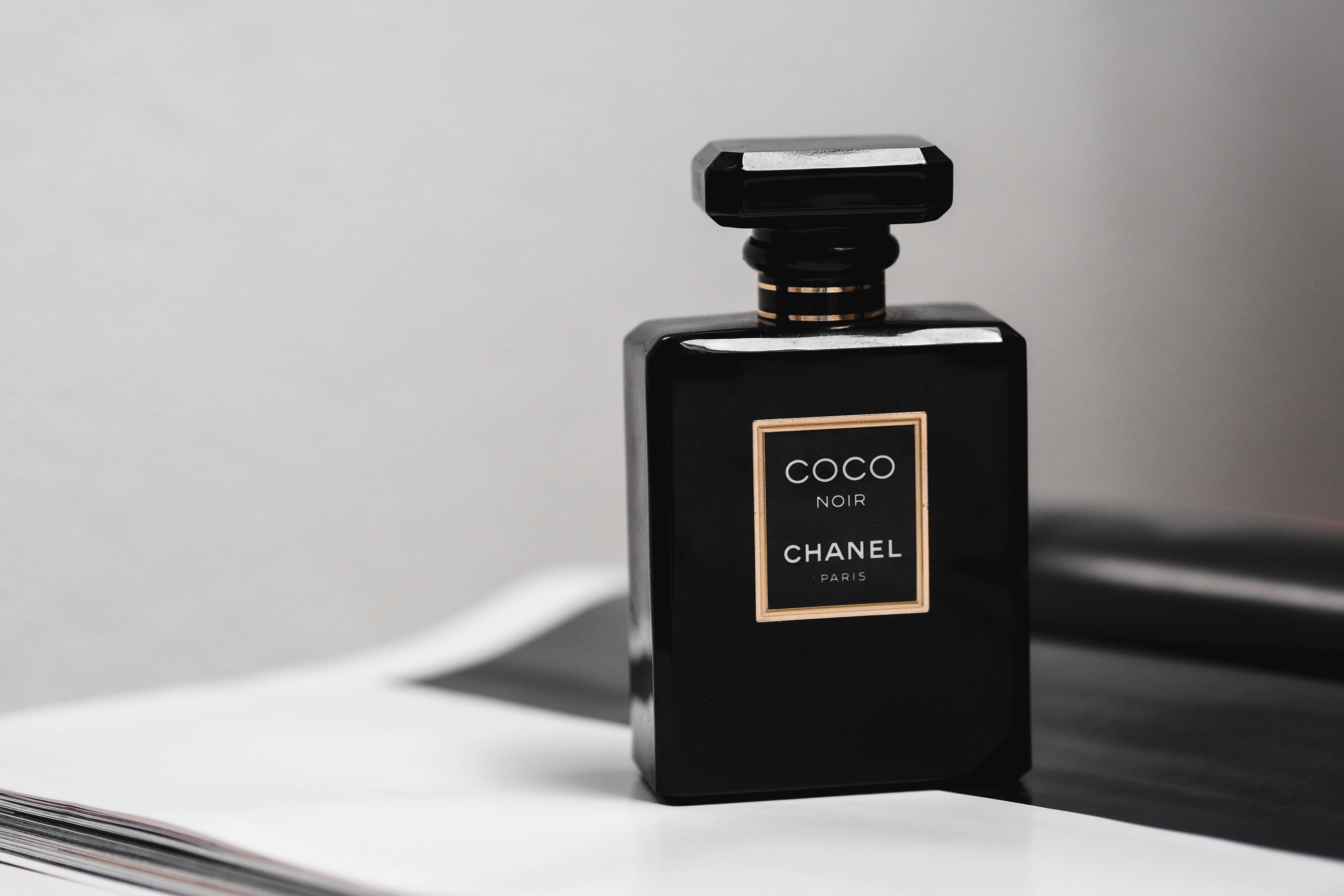 香奈兒Coco Noir香水 壁紙 — 一個時尚的黑色 Chanel Coco Noir 香水瓶，帶有金色點綴，優雅地放置在白色表面上，背景為柔和的灰色。