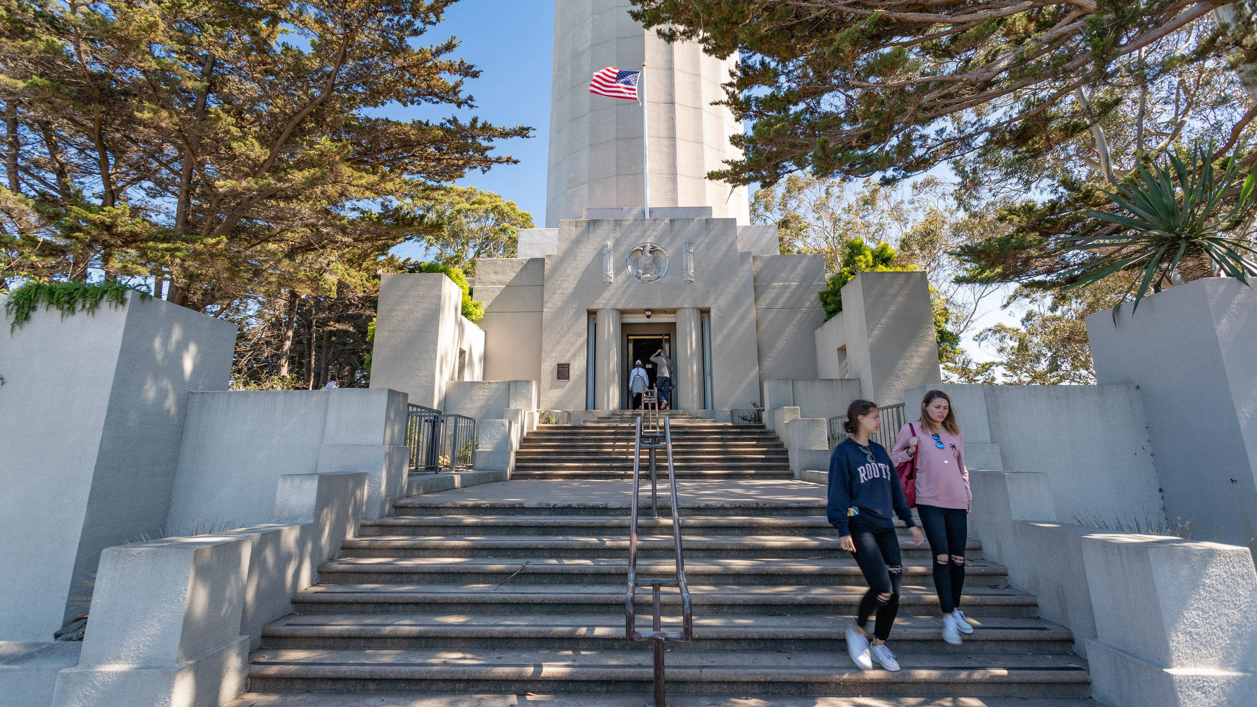 Coit Tower階梯步道 壁紙 — 兩個女孩沿著石階走向一座高大的白色紀念碑，頂端有美國國旗，周圍是高聳的樹木，陽光明媚。