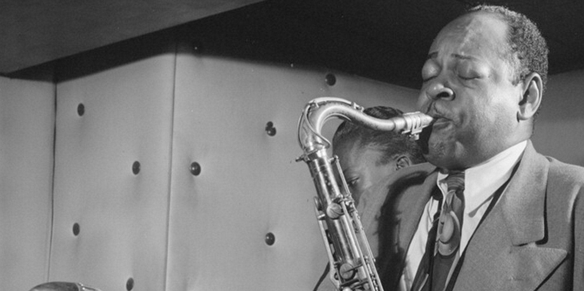 Coleman Hawkins 演奏薩克斯風 壁紙 — 一位爵士音樂家在昏暗的房間裡熱情地吹奏色士風，房間有簇絨牆壁。