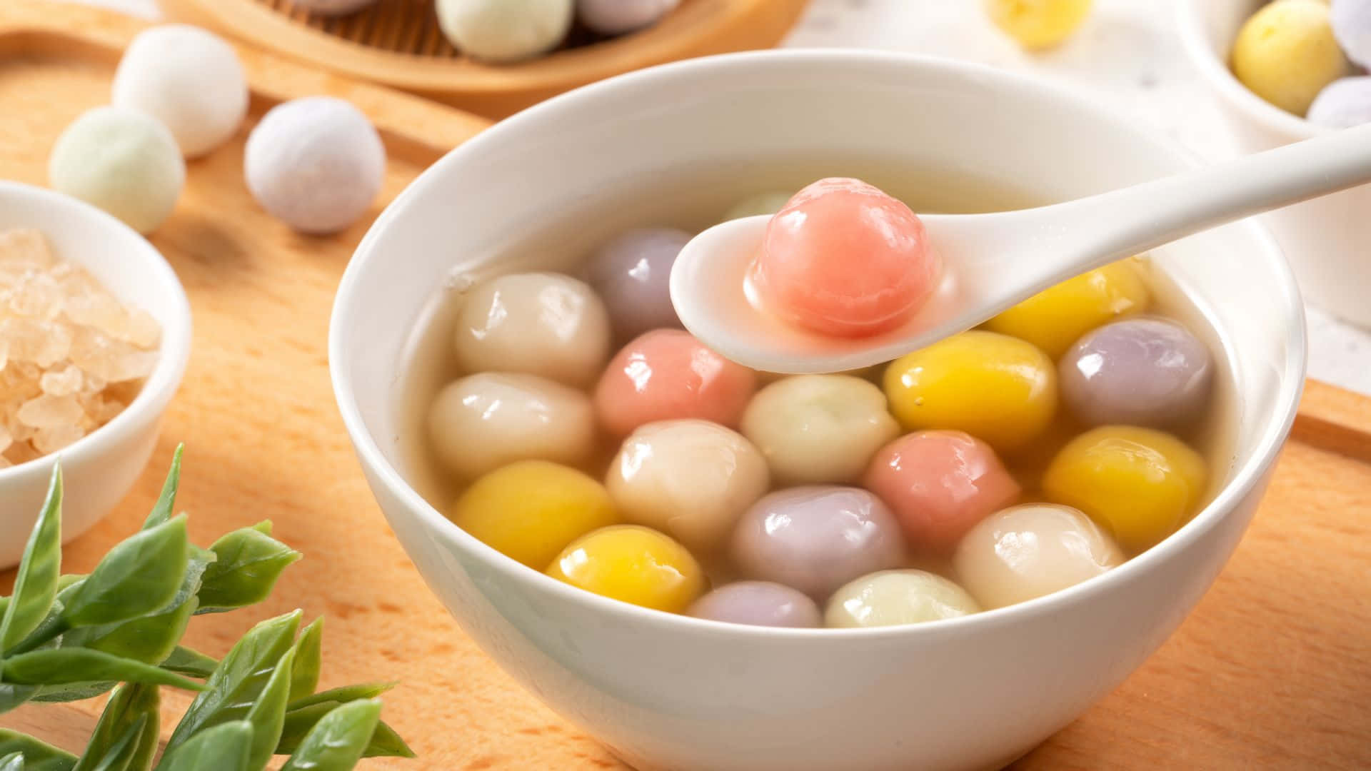  壁紙 — 一個白色碗裡裝滿了色彩繽紛的半透明湯圓，浸泡在甜湯中，湯匙正舀起一個粉紅色的湯圓。