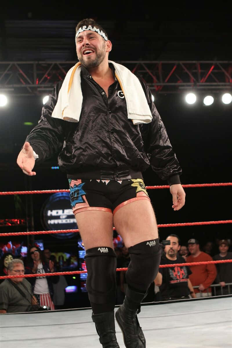 Colt Cabana摔角擂台入場 壁紙 — 一名身穿黑色裝備和白色毛巾的摔角手在明亮的場館燈光下於擂台上慶祝。