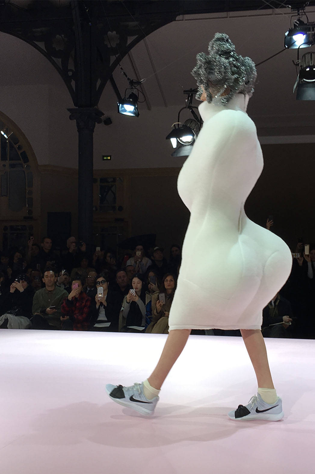Comme Des Garçons 膨脹身形連衣裙 壁紙 — 一位模特兒穿著白色雕塑緊身衣和運動鞋走在粉紅色天橋上，觀眾捕捉這一刻。
