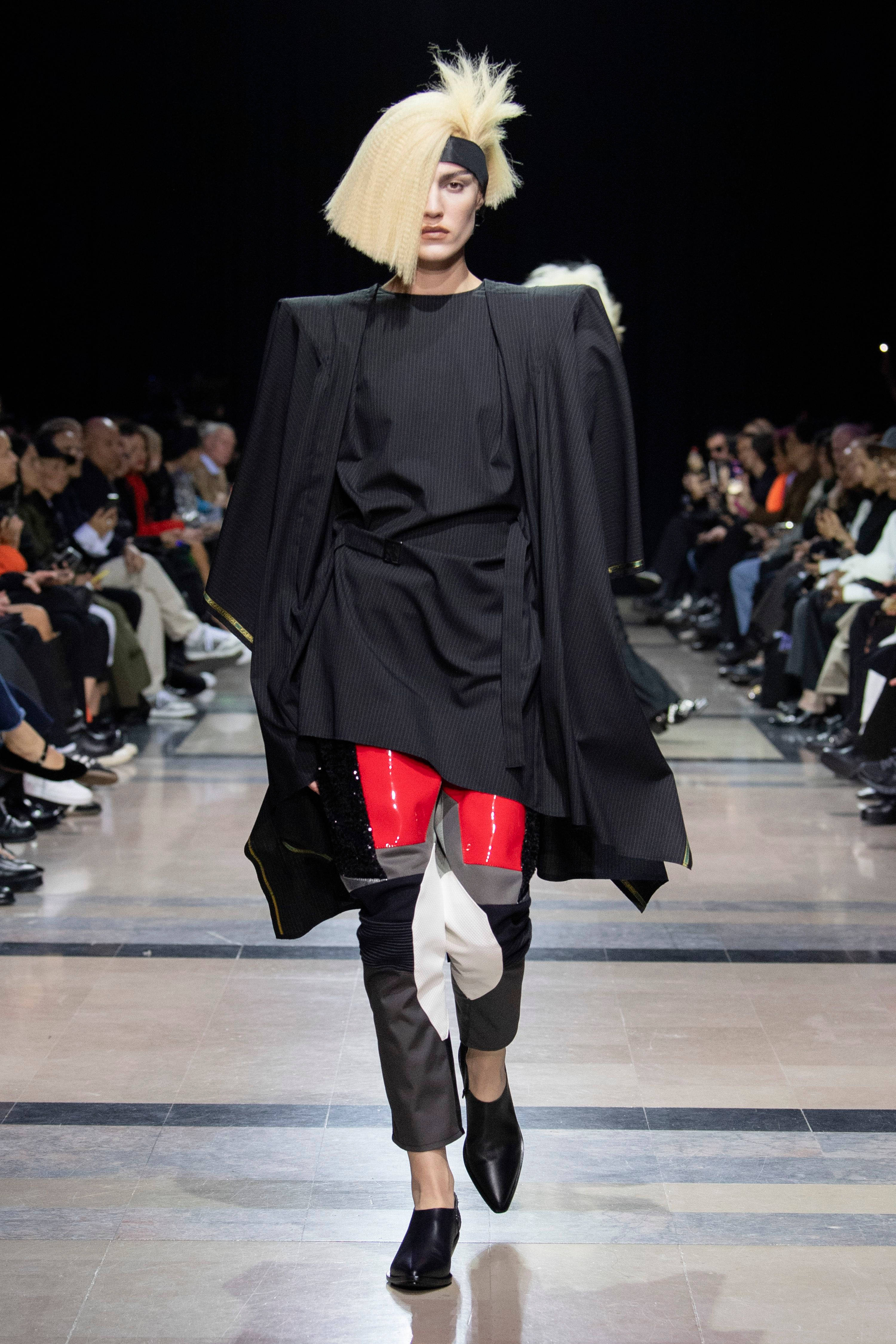 Comme Des Garçons 黑色造型服裝 壁紙 — 一位模特兒留著大膽的金色鮑伯頭，戴著黑色頭帶，身穿前衛黑色細條紋服裝，搭配紅色和白色點綴，走在天橋上。