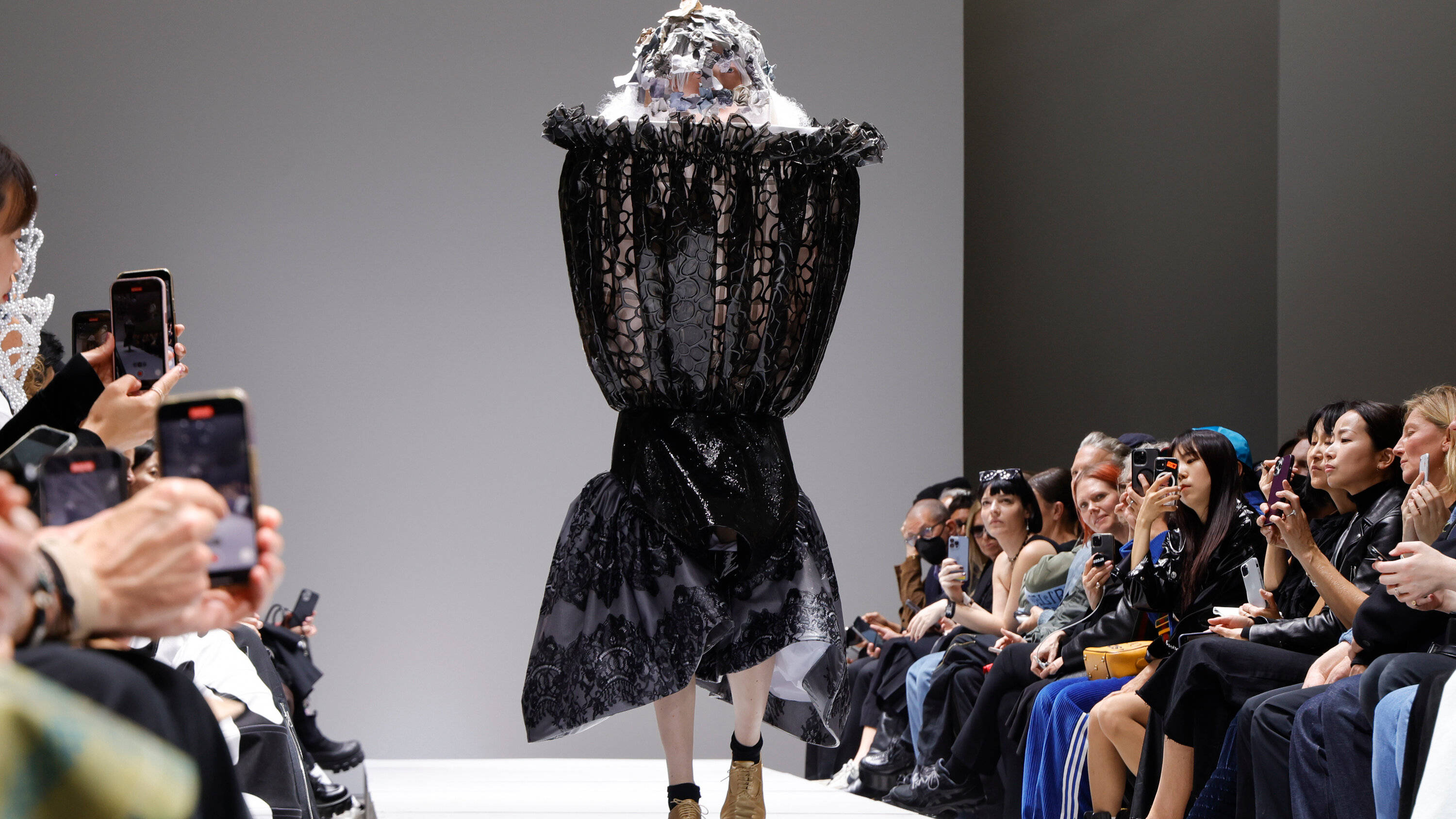 Comme Des Garçons 2023 春季系列 壁紙 — 模特兒穿著前衛黑色蕾絲連衣裙和雕塑頭飾走秀，觀眾用手機捕捉這一刻。