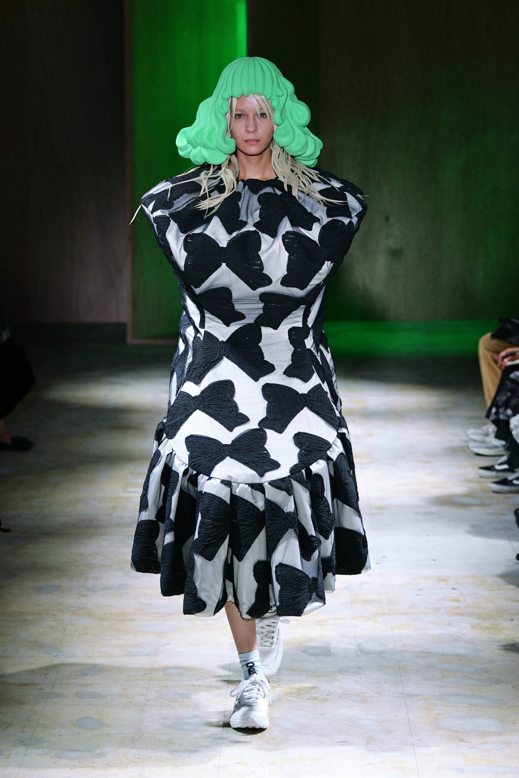 Comme Des Garçons 黑白絲帶無袖連衣裙 壁紙 — 模特兒穿著大膽的黑白蝴蝶結圖案連衣裙，搭配亮綠色假髮和白色運動鞋走在天橋上。
