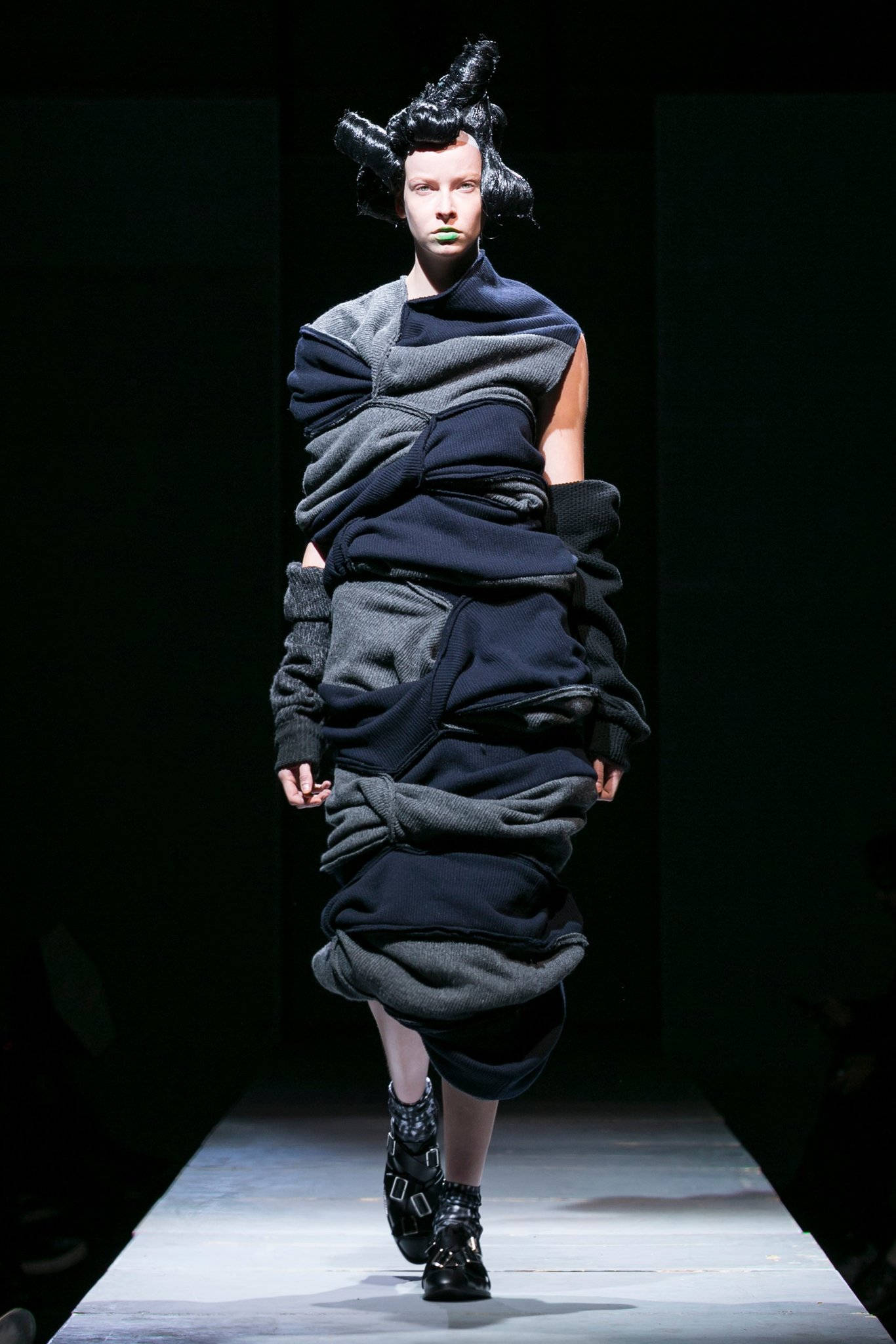 Comme Des Garçons 深色拼布連衣裙 壁紙 — 模特兒在戲劇性的舞台燈光下，穿著前衛的層疊黑色和灰色服裝走秀。