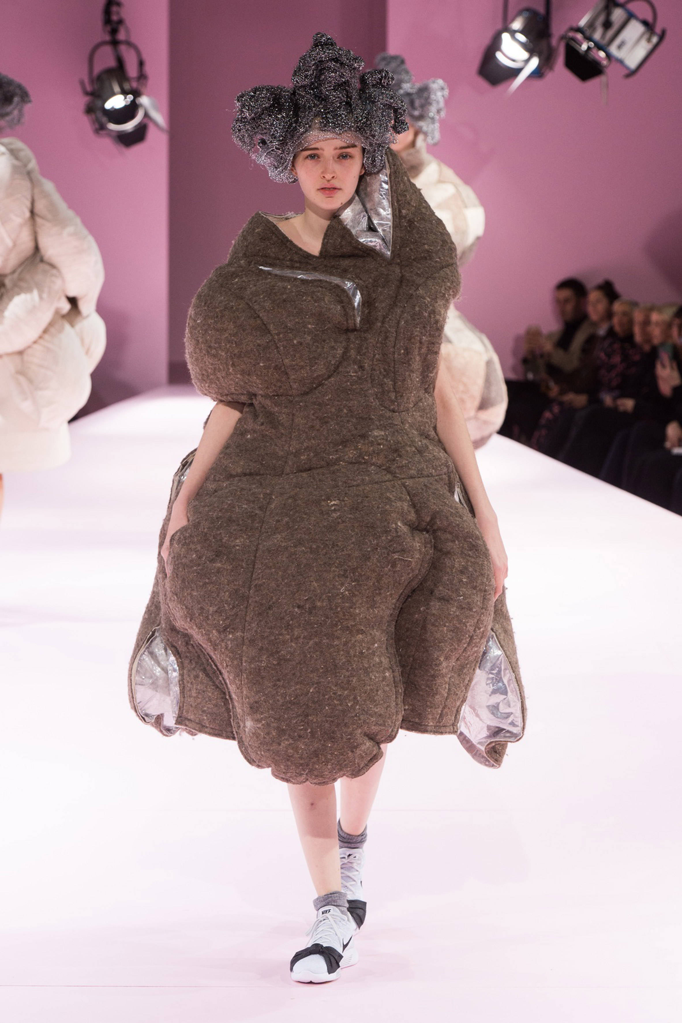 Comme Des Garçons 巨大棕色大衣 壁紙 — 模特兒在粉紅色燈光的時裝天橋上走秀，身穿帶有銀色點綴的豐盈棕色質感服裝和雕塑頭飾。