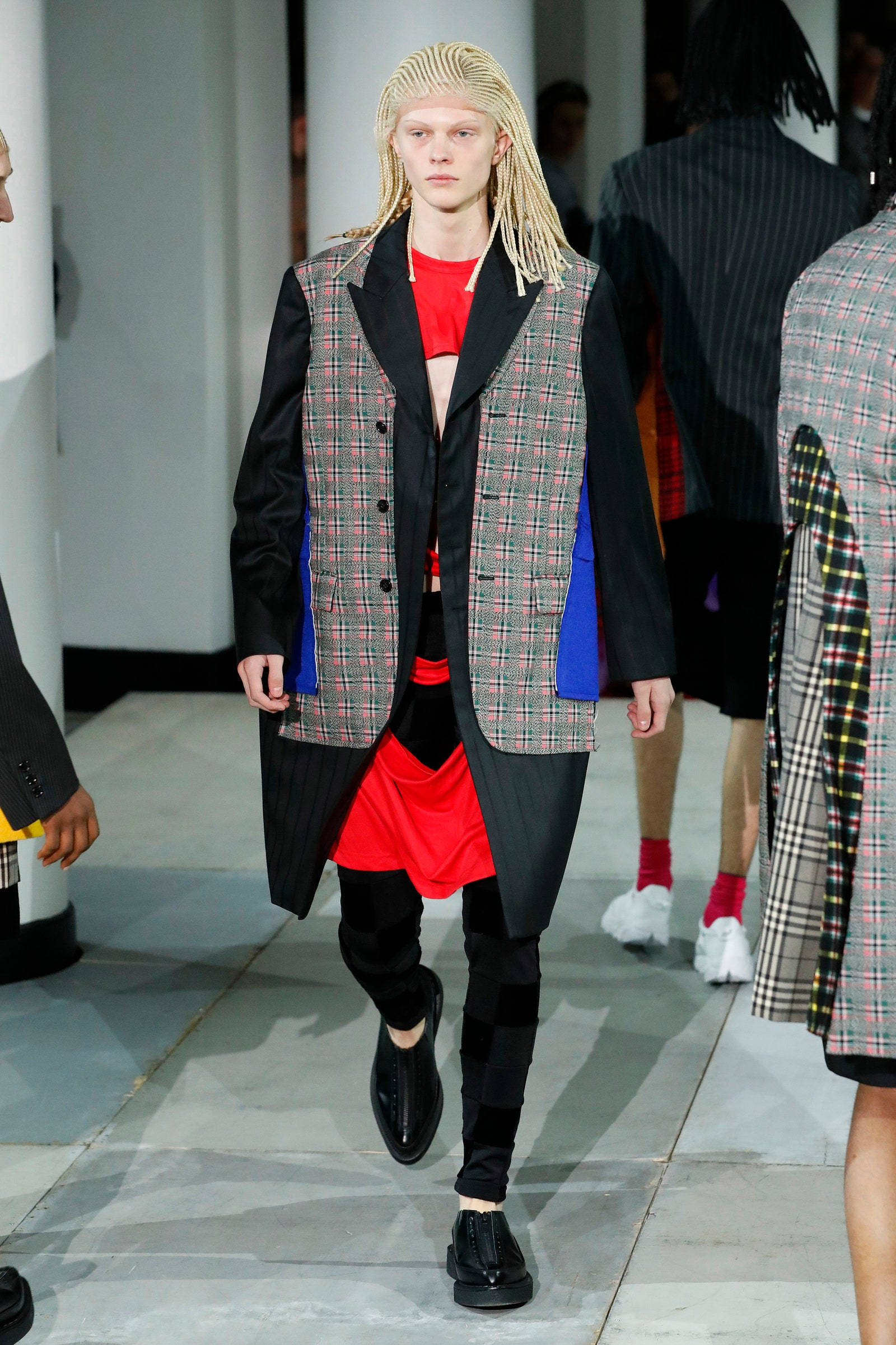 Comme des Garçons 格紋背心 壁紙 — 一位金髮辮子模特兒穿著大膽的格紋背心，內搭紅色上衣和黑色褲子走秀。