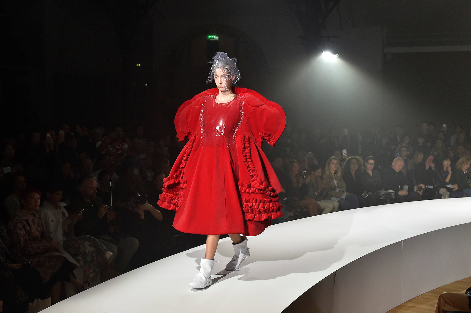 Comme Des Garçons 紅色蓬袖連衣裙 壁紙 — 一位模特兒穿著寬大的紅色荷葉邊連衣裙，在聚光燈下自信地走在彎曲的T台上。