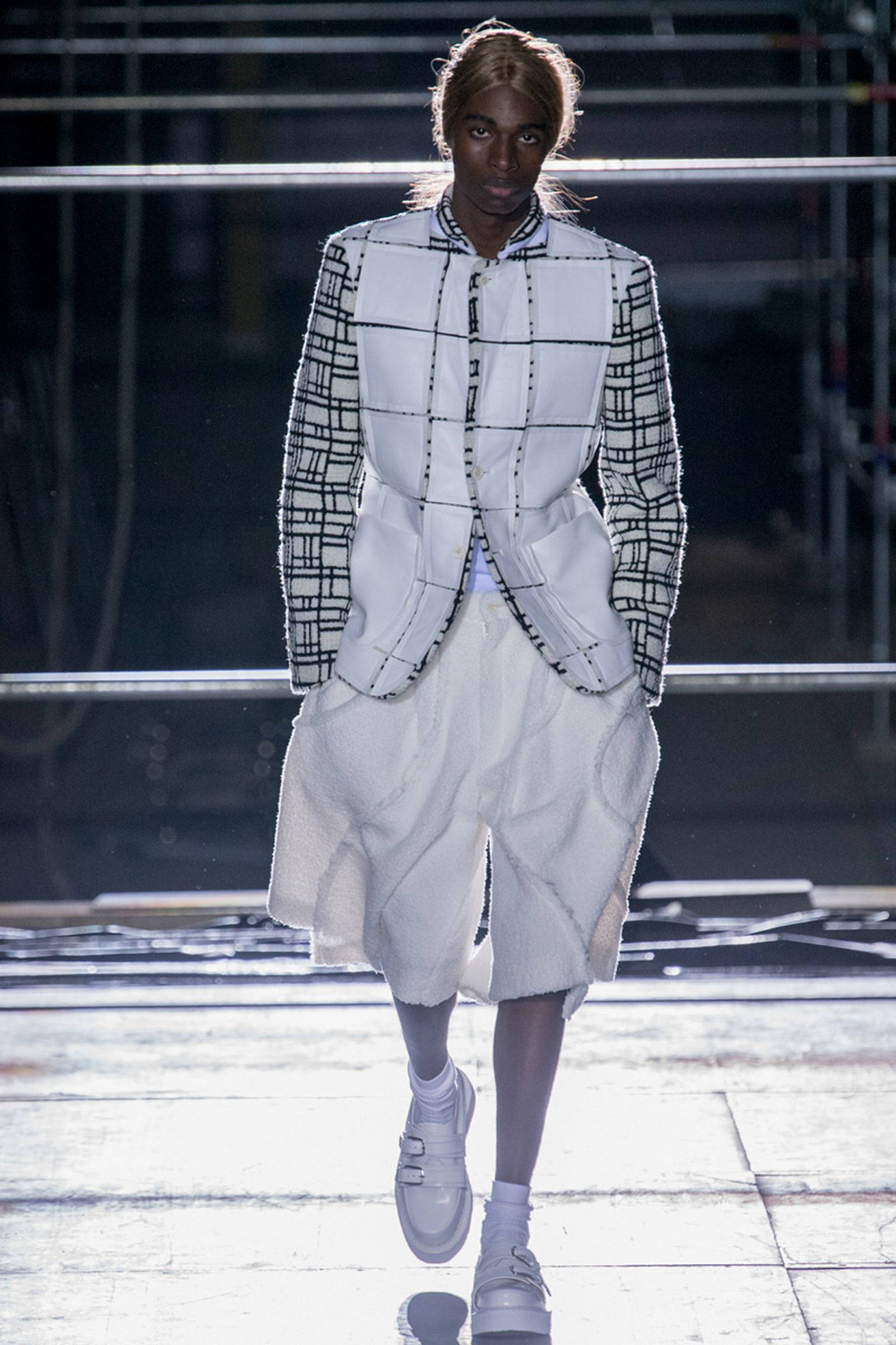 Comme Des Garçons 黑白格紋服裝 壁紙 — 模特兒自信地走在天橋上，身穿大膽的黑白幾何西裝外套，搭配白色垂墜短褲和厚底白色運動鞋。