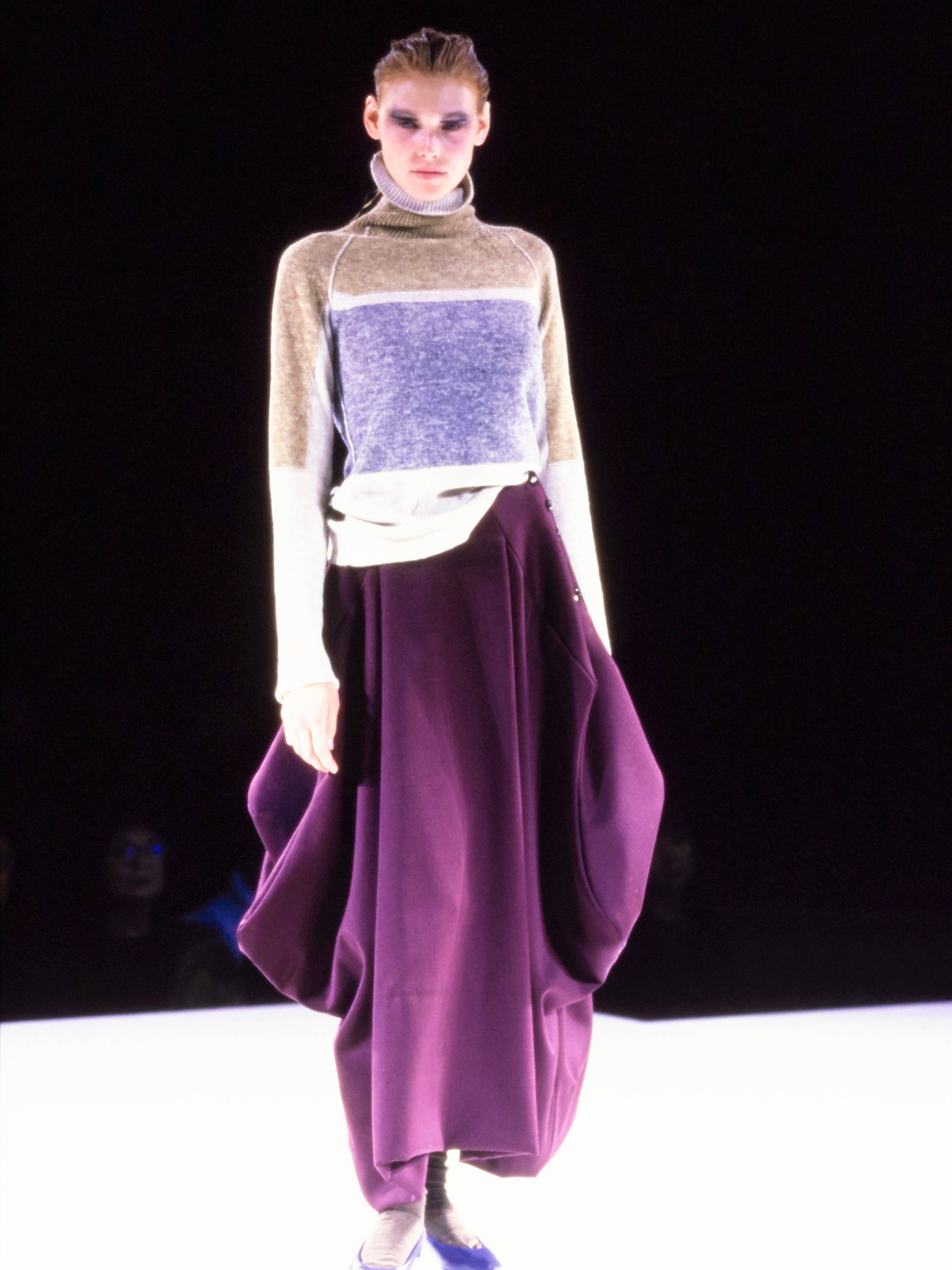 Comme Des Garçons 棕紫配色服裝 壁紙 — 一位模特兒自信地走在天橋上，穿著分層造型，包括一件色塊毛衣和一條飄逸的紫色裙子，在明亮的舞台燈光下。