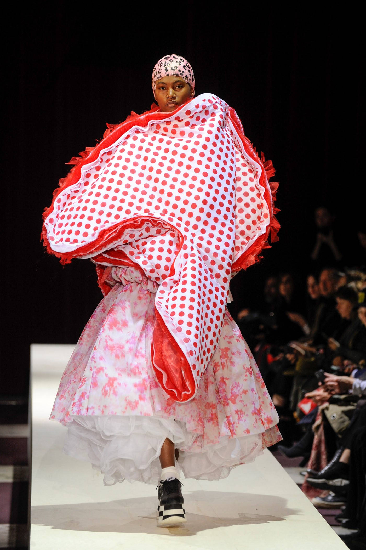 Comme Des Garçons 粉白花卉連衣裙配紅白波點斗篷 壁紙 — 一位模特兒自信地走在天橋上，身穿大膽的紅白波點套裝，搭配花卉裙和頭巾。