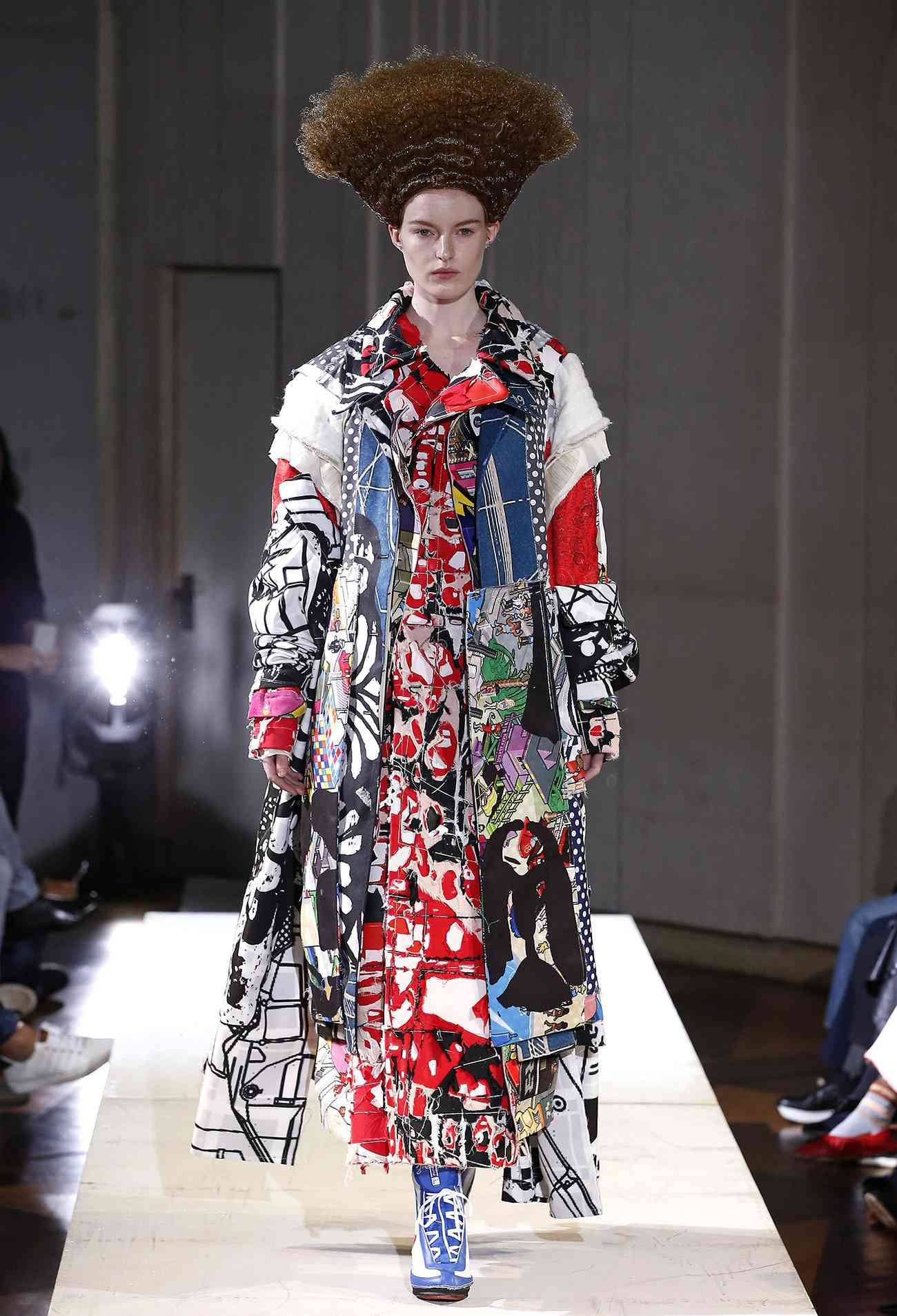 Comme Des Garçons 大膽拼接外套 壁紙 — 一位模特兒在明亮的舞台燈光下，穿著帶有圖案印花的搶眼拼布外套和高聳的非洲式假髮，走在天橋上。