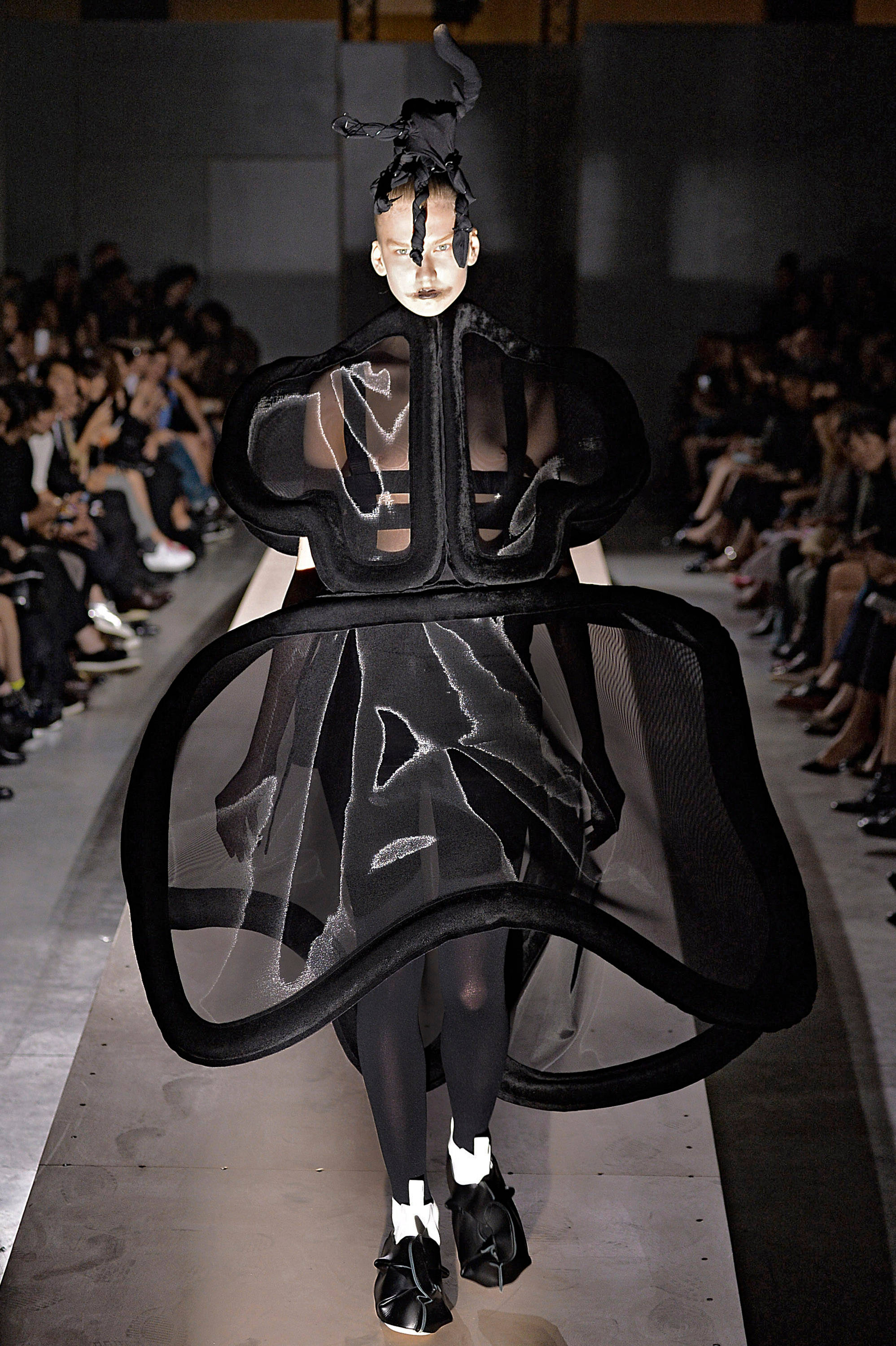 Comme Des Garçons 春季2014成衣系列 壁紙 — 一位模特兒在明亮的燈光下，穿著帶有雕塑層次和戲劇性頭飾的前衛黑色連衣裙走秀。