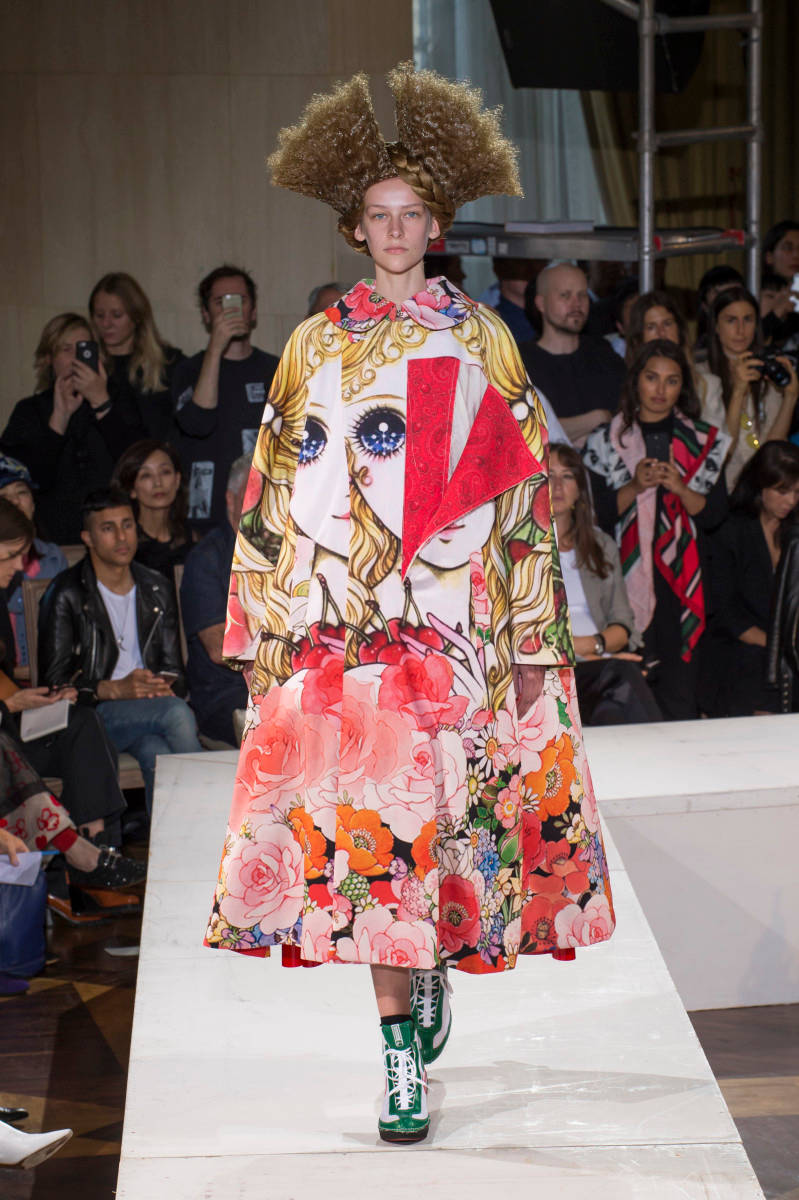 Comme Des Garçons 紅色娃娃花紋連衣裙 壁紙 — 一位模特兒穿著充滿活力的花卉和人臉印花長袍、高聳的非洲式假髮和綠色靴子走在天橋上。