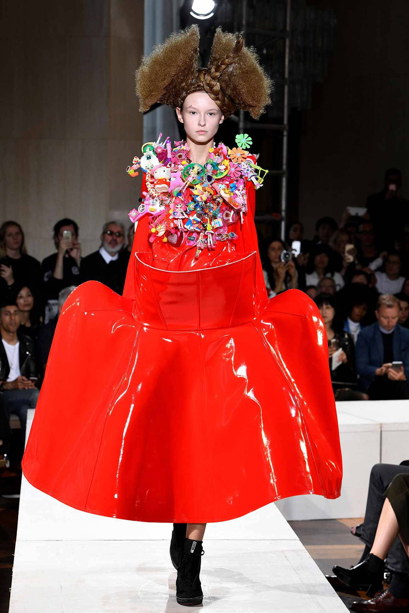 Comme Des Garçons 紅色皮革不規則連衣裙 壁紙 — 一位模特兒在時裝天橋上走秀，身穿大膽的紅色亮面連衣裙，飾有色彩繽紛的毛絨玩具和一頂大的棕色非洲式假髮。
