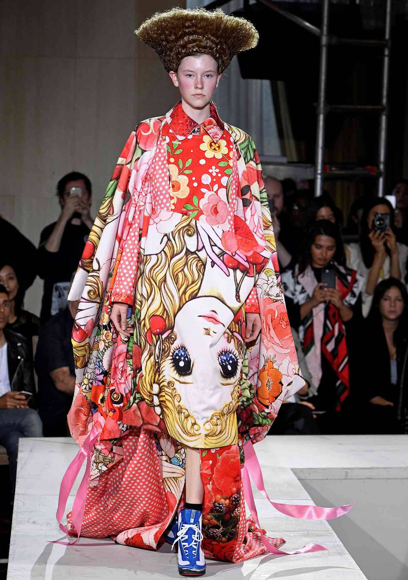 Comme Des Garçons 紅色娃娃臉連衣裙 壁紙 — 一位模特兒在明亮的燈光下，穿著一件帶有大型臉部印花的鮮豔花卉和服，戴著高聳的棕色假髮，走在天橋上。