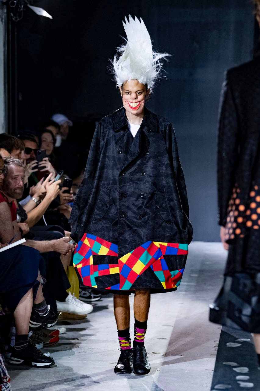 Comme Des Garçons微笑面具造型 壁紙 — 一位模特兒戴著白色羽毛頭飾和小丑妝容，穿著黑色外套和彩色幾何裙走秀。