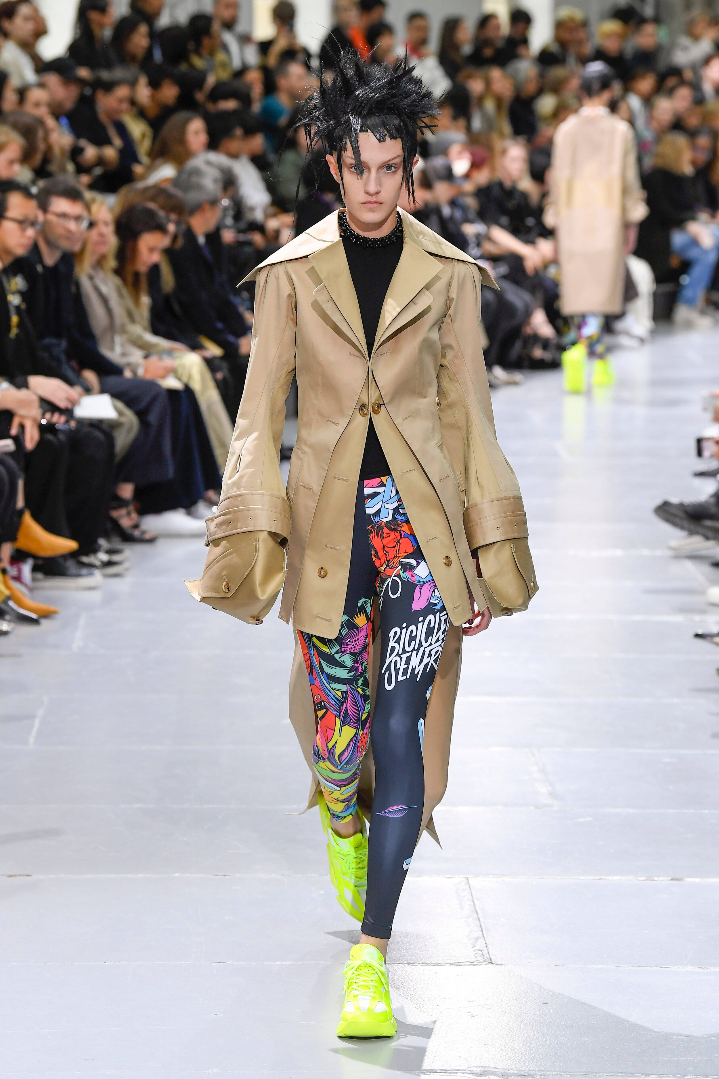 Comme Des Garçons 西裝外套與印花褲 壁紙 — 一位模特兒身穿米色風衣，搭配鮮豔的塗鴉印花緊身褲和螢光綠色運動鞋，在T台上走秀。