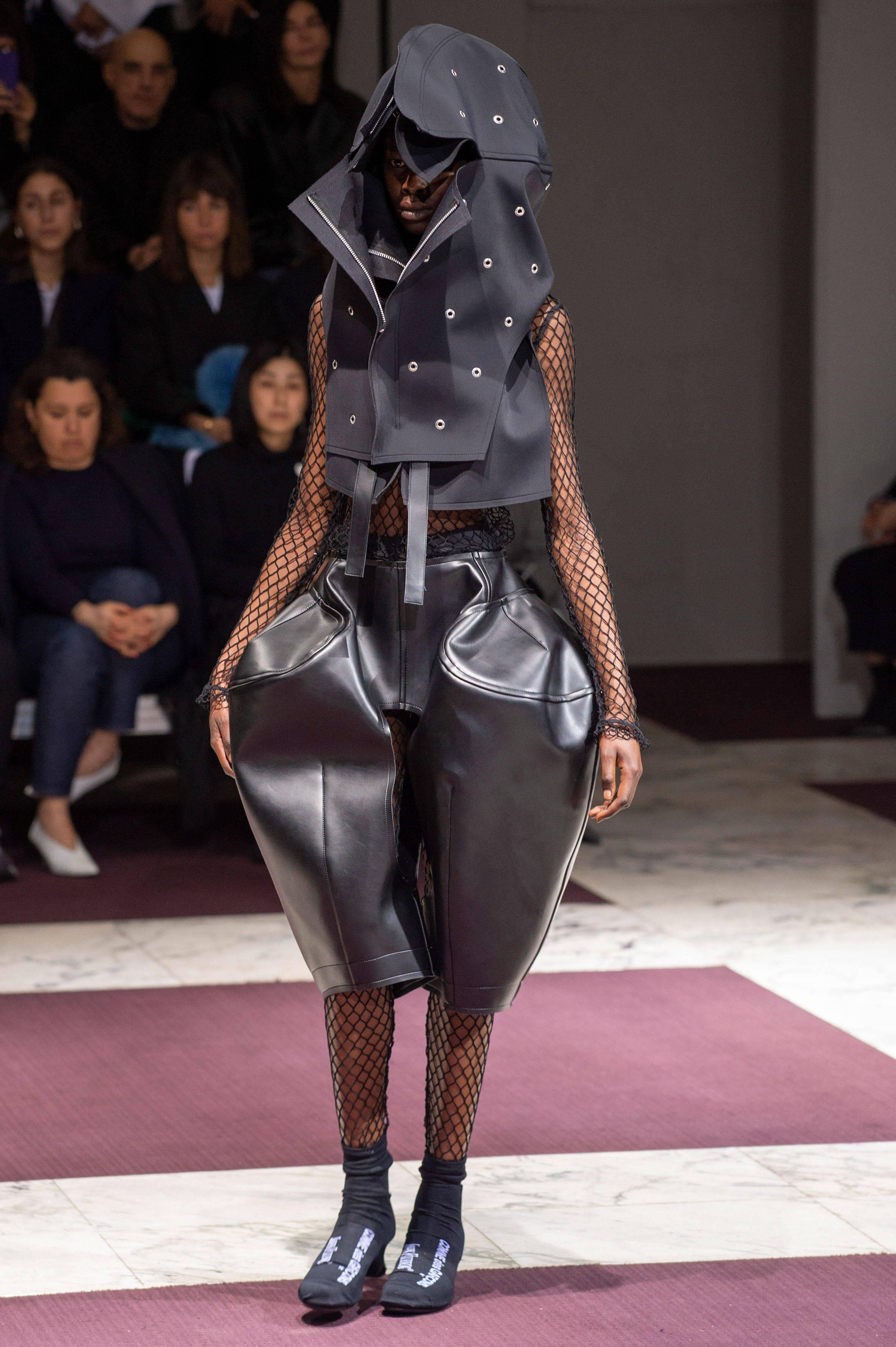 Comme Des Garçons 全黑服裝 壁紙 — 一位模特兒穿著前衛黑色皮革服裝，搭配漁網袖和超大連帽，在時裝展台上行走。