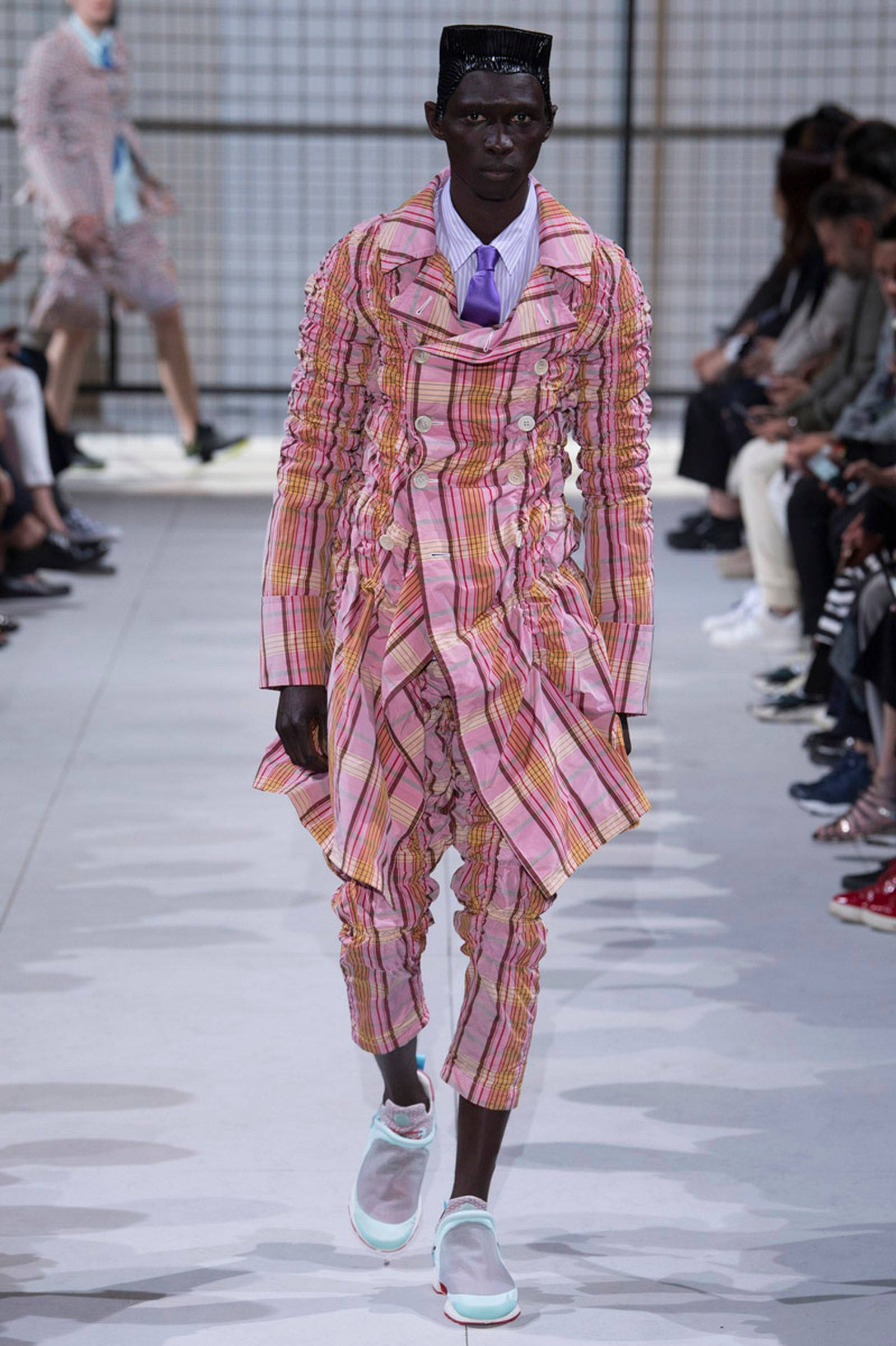 Comme Des Garçons 橙粉格紋服裝 壁紙 — 一位模特兒自信地走在天橋上，身穿寬鬆的粉紅色格紋西裝，帶有荷葉邊細節和淺藍色運動鞋。