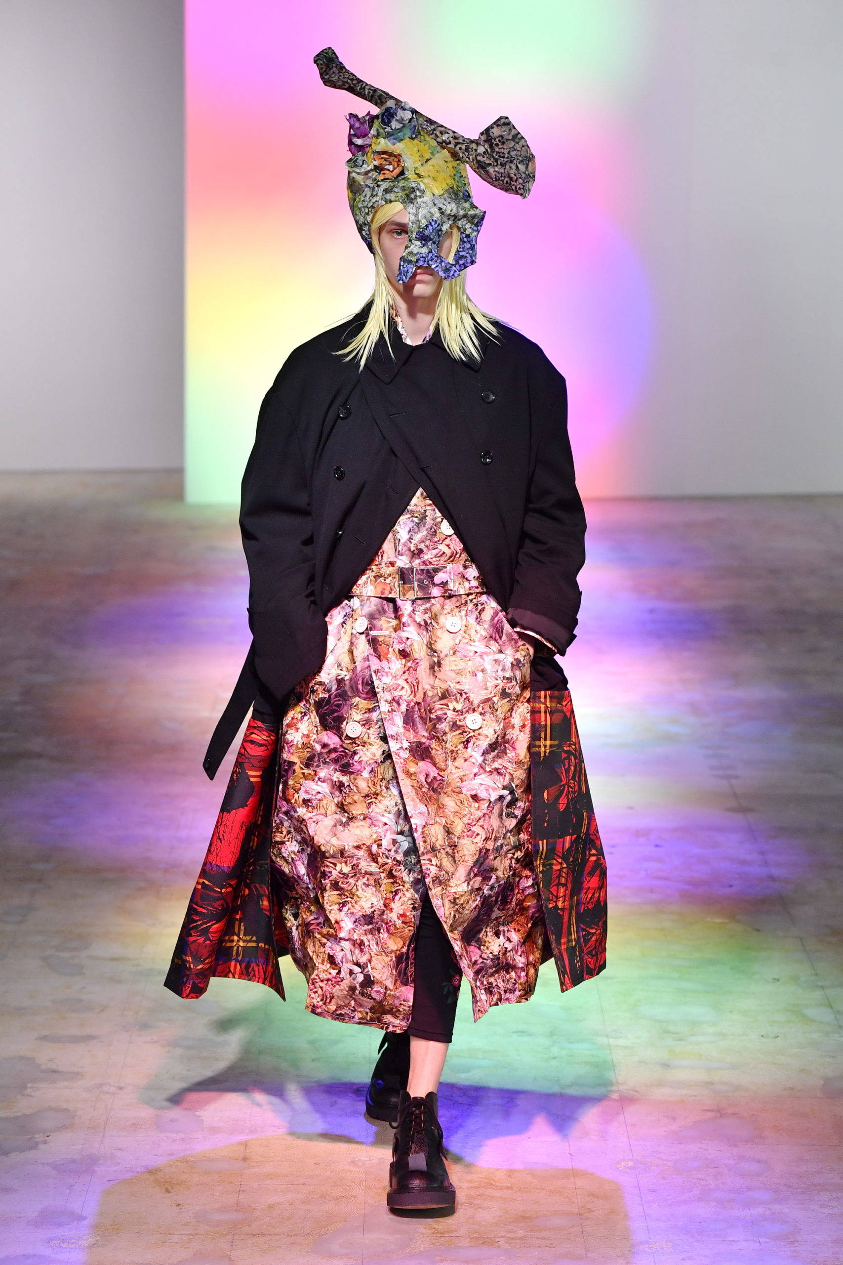 Comme Des Garçons 花紋長裙配外套 壁紙 — 一位模特兒在時裝展台上行走，穿著黑色外套配搭花卉圖案裙子，並在充滿活力的舞台燈光下戴著色彩繽紛的圖案口罩。